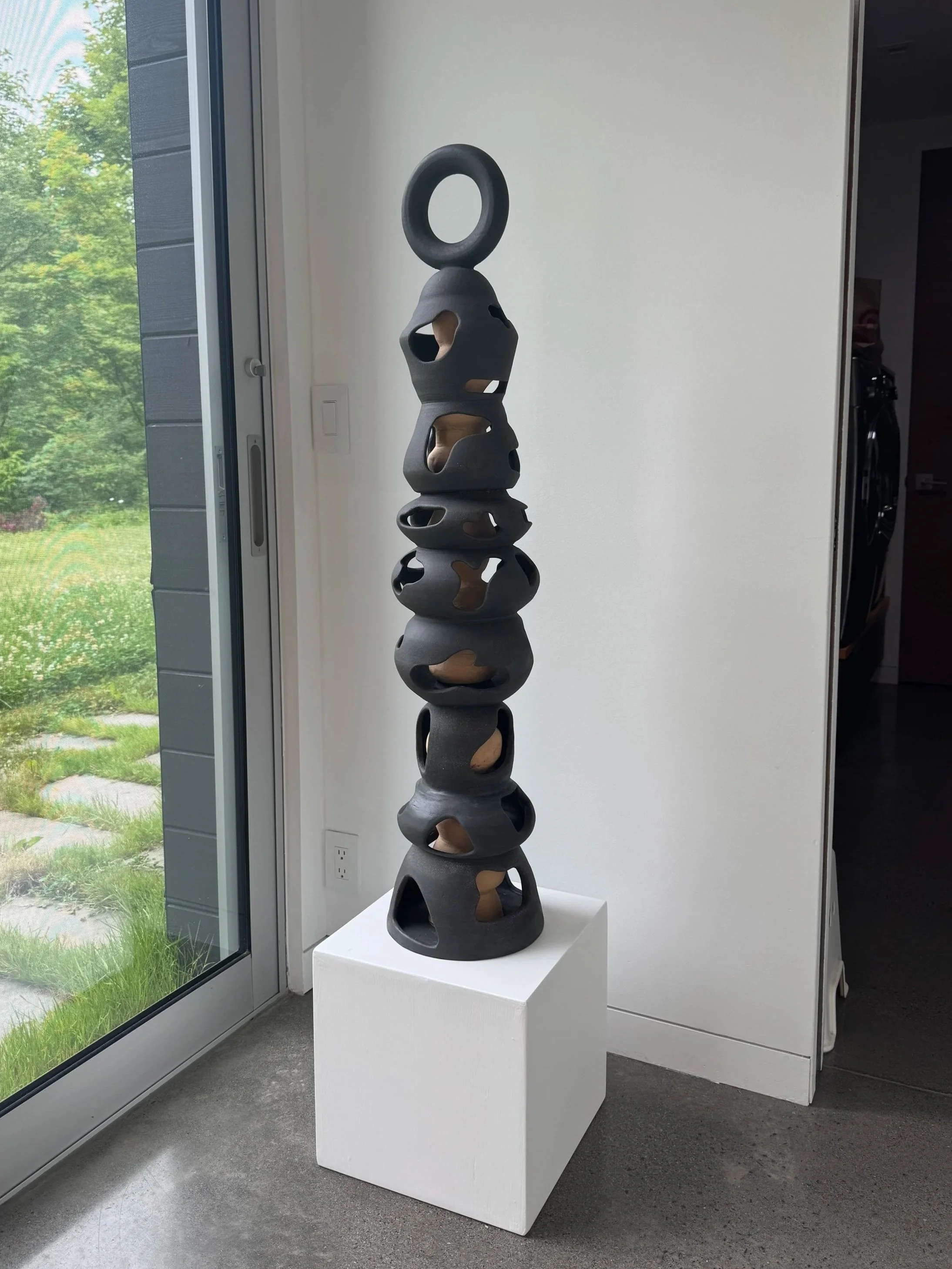 A tall, black abstract sculpture with multiple hollow, rounded sections stacked vertically, topped with a large loop, standing on a white pedestal.