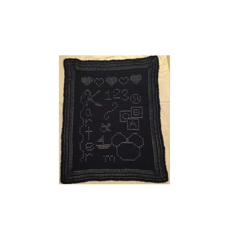 A black crocheted or knitted blanket with various white or light-colored patterns and symbols, including hearts, numbers, and other decorative shapes.