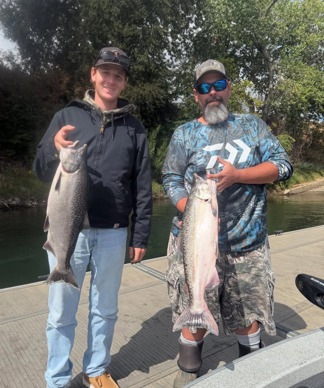 Two men on a dock holding large fish, smiling, with trees and water in the background.