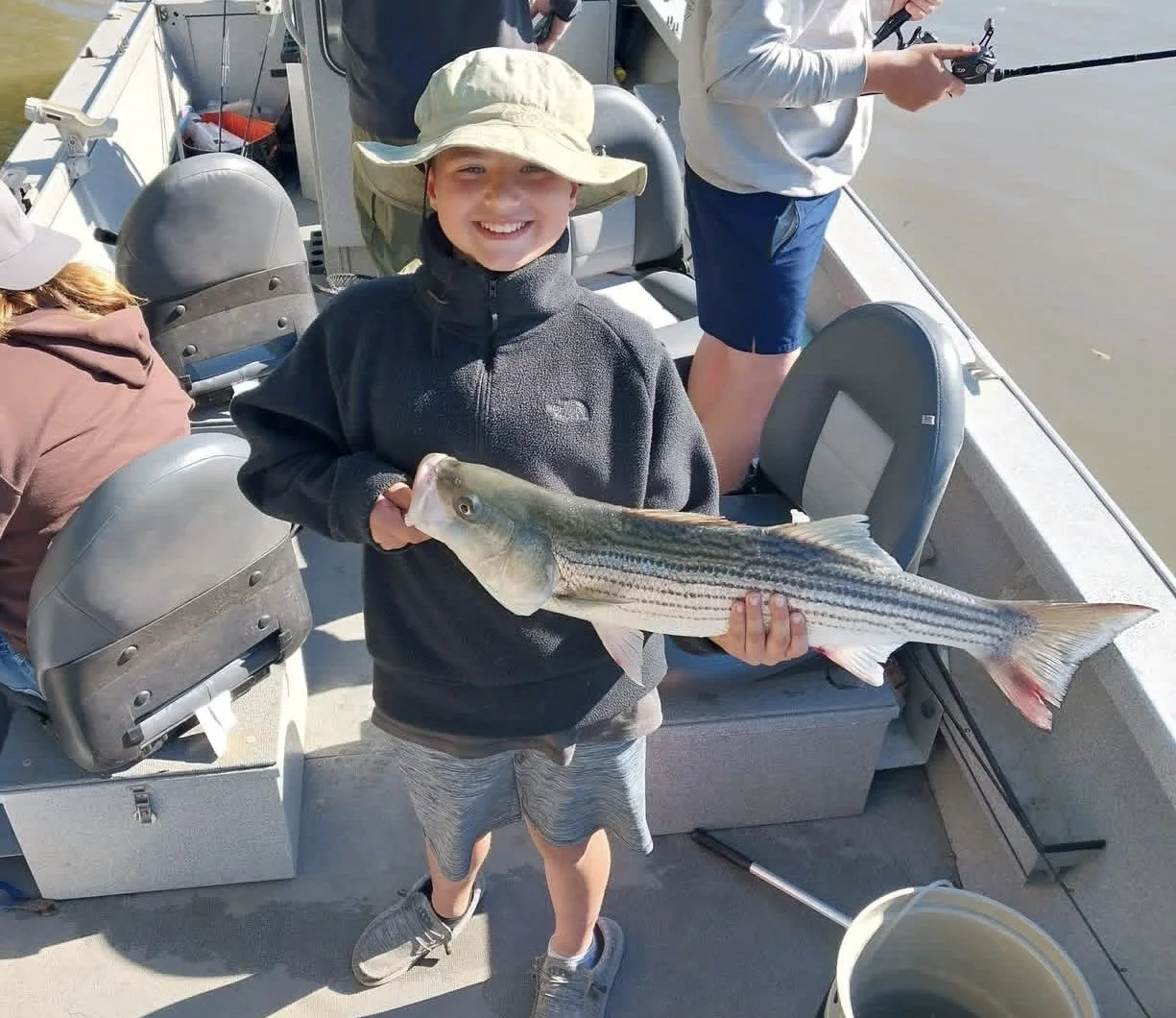A young boy on a boat holding a large striped bass fish, smiling at the camera, wearing a beige sun hat and dark sweatshirt with other people fishing in the background.