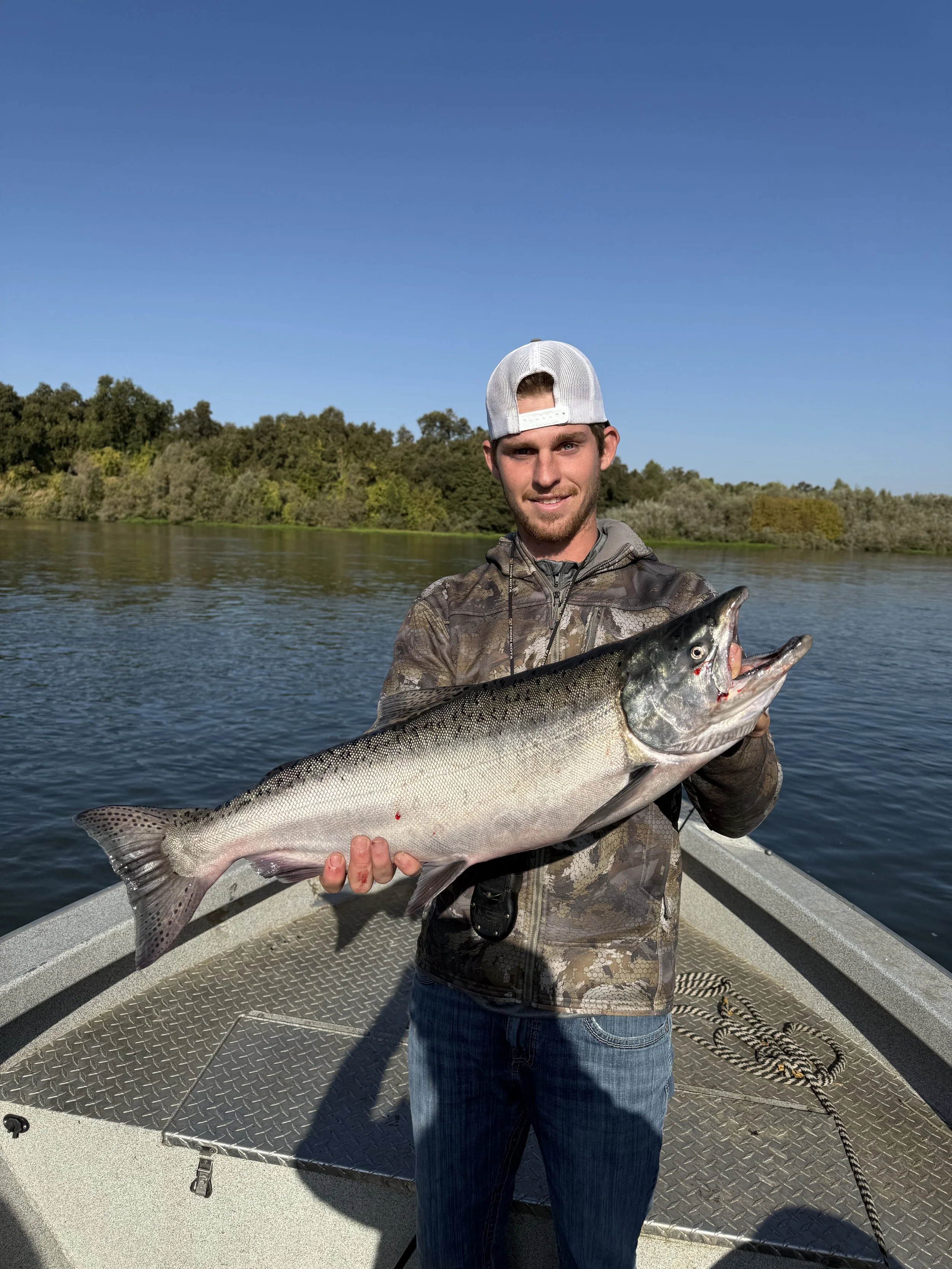 A young man in camouflage jacket and baseball cap holding a large fish on a boat in a lake with trees in the background.