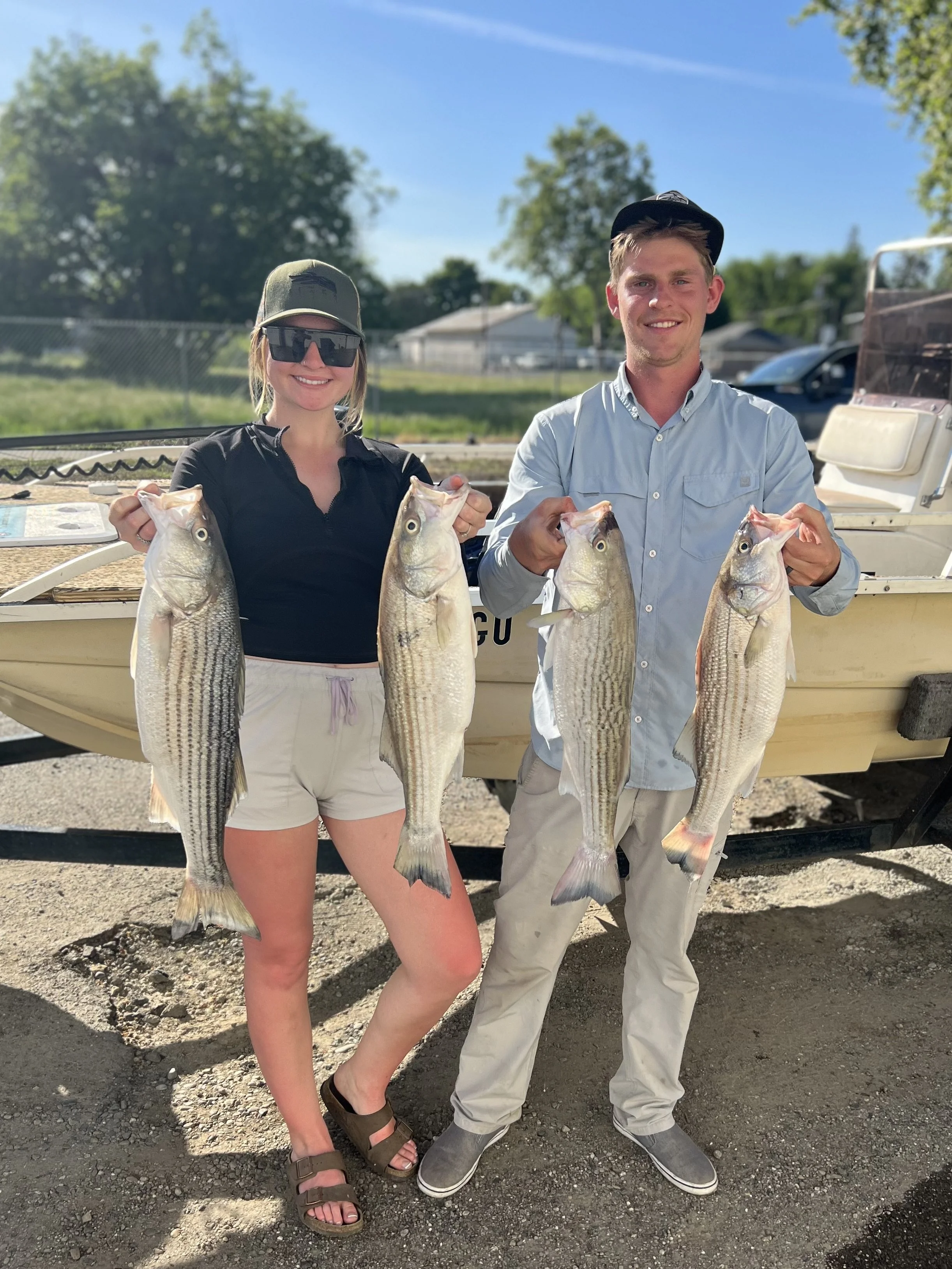 A woman and a man standing outdoors in front of a boat, each holding two large fish, smiling. The woman is wearing a black shirt, white shorts, sunglasses, and sandals; the man is wearing a light blue shirt, beige pants, and a black cap.