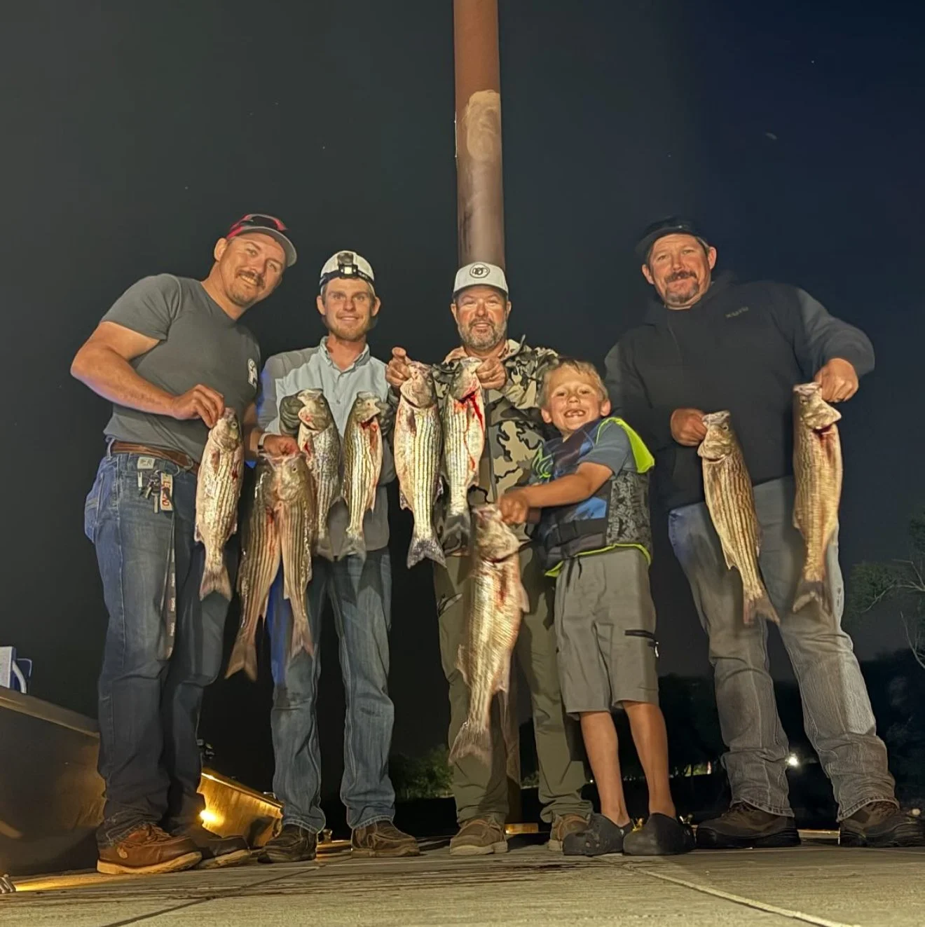 A group of five men and one boy are standing together outdoors at night, holding up large fish they have caught during a fishing trip.
