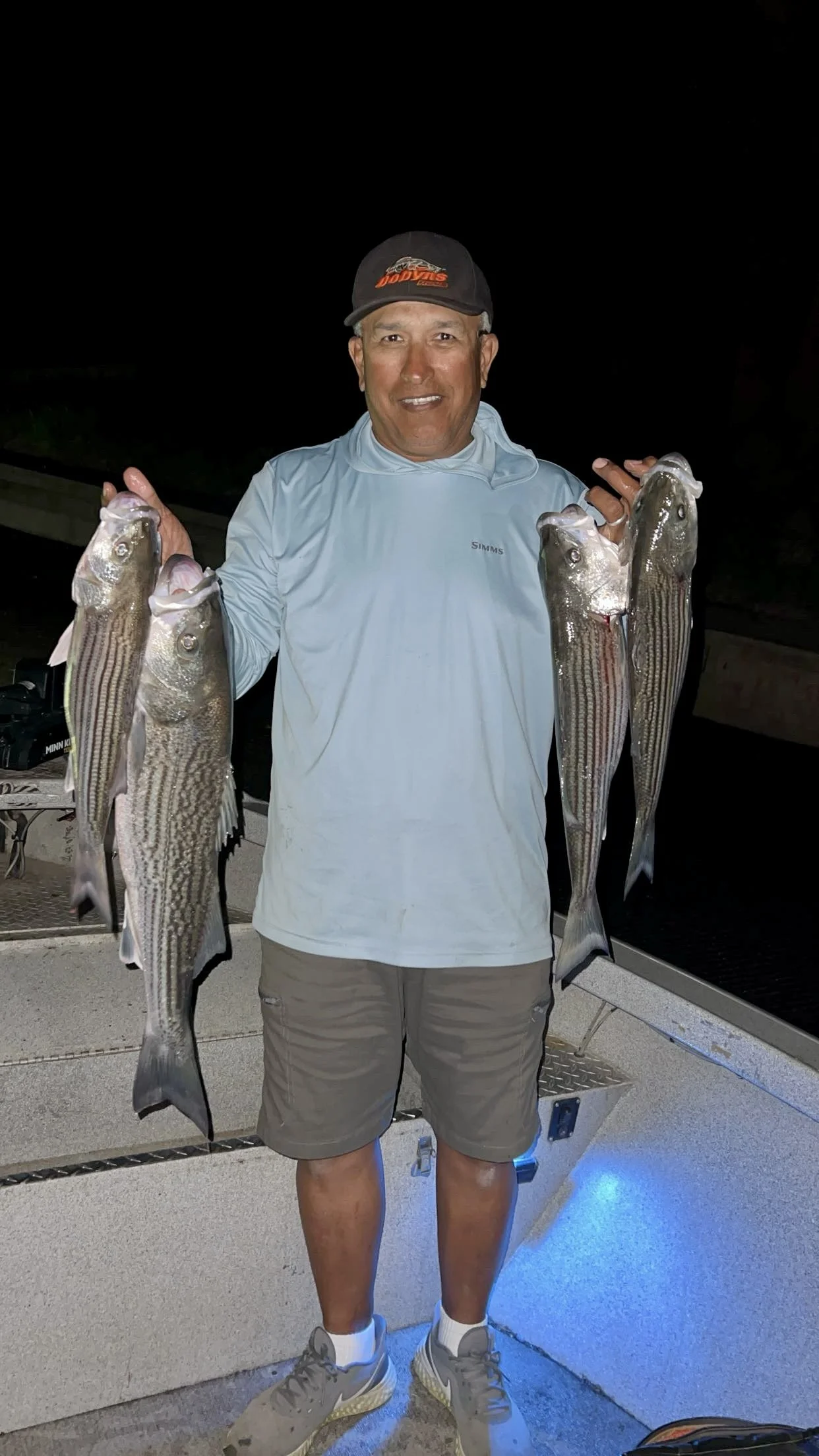 A man standing on a boat deck at night, smiling and holding four large striped fish, two in each hand.