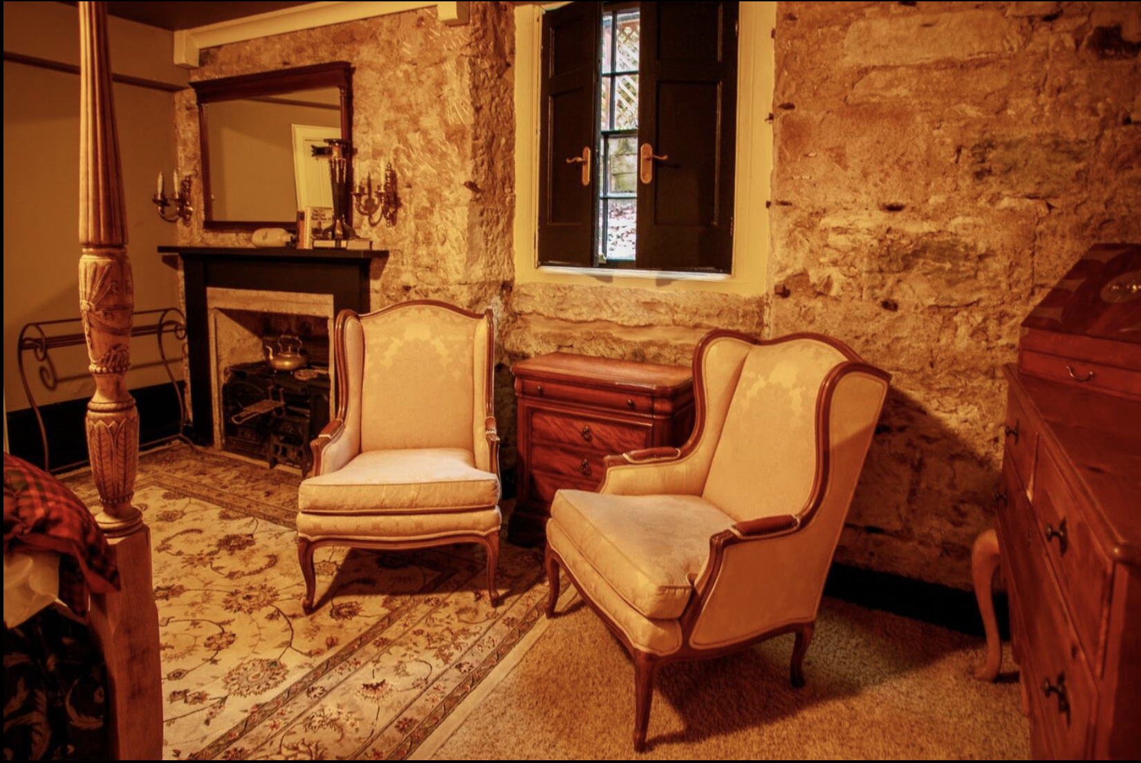 A cozy vintage-style room with two plush beige armchairs, a small wooden chest, a stone wall, a window with open dark shutters, a fireplace with a mirror and decorative candle holders, and floral patterned rug.