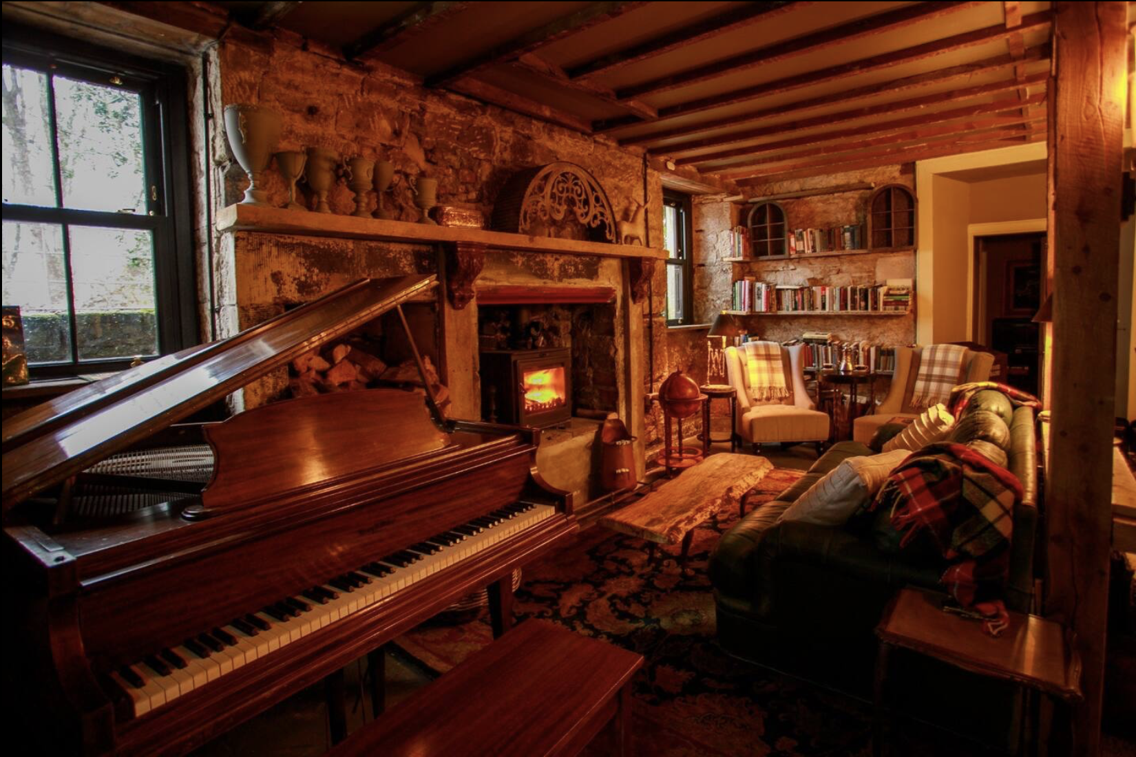 Cozy living room with a grand piano, stone fireplace with a fire, wooden ceiling, and bookshelf. Comfortable armchairs and sofa with plaid blankets, decorated for a warm, inviting atmosphere.
