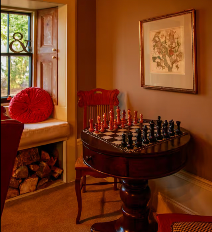 A cozy corner with a window seat, a round red pillow, and a stack of firewood beneath a window. A vintage wooden table with a chessboard set up, a leafy framed artwork on the wall, and a wooden chair.