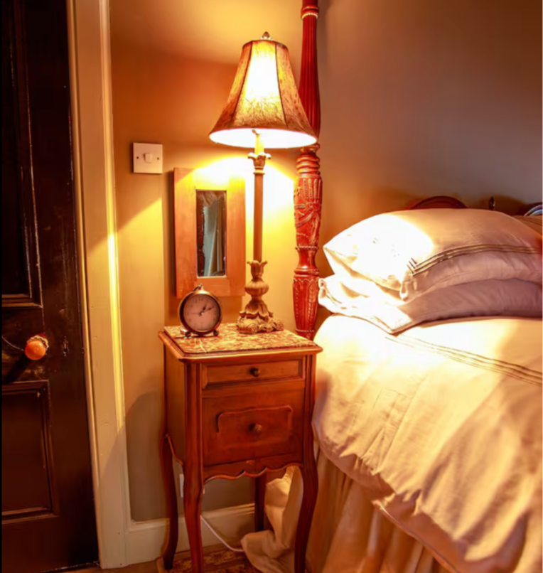 A bedroom nightstand with a tall lamp, an alarm clock, and a small mirror. Part of a bed with pillows is visible to the right.