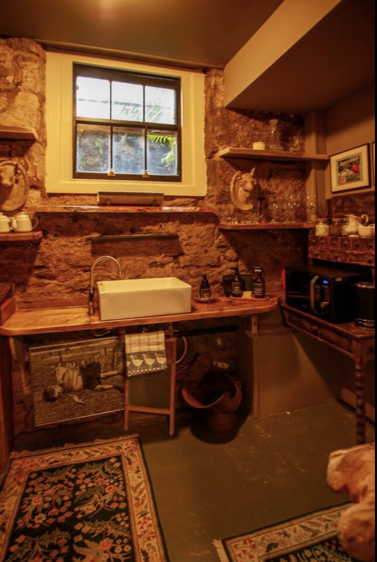 Rustic kitchen with stone walls, a window, a farmhouse sink, wooden shelves with decorative items, a small microwave, and a colorful area rug.
