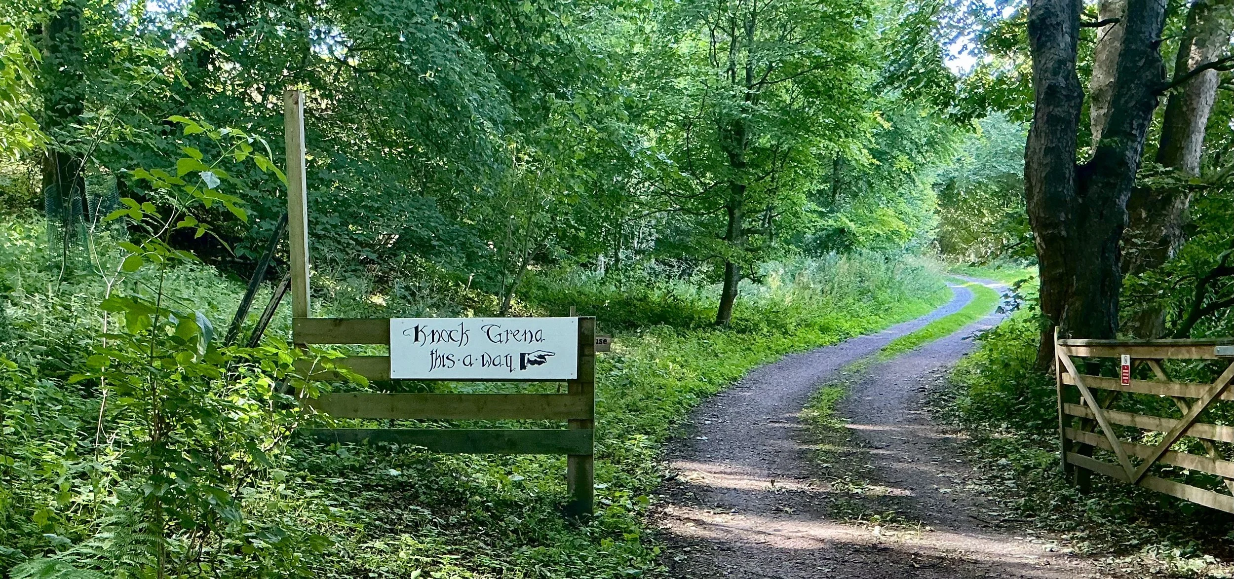 A dirt pathway through a green wooded area with a sign that reads 'Knock Grena This a way' on the left side, and a small wooden gate on the right side. Trees and foliage surround the scene.