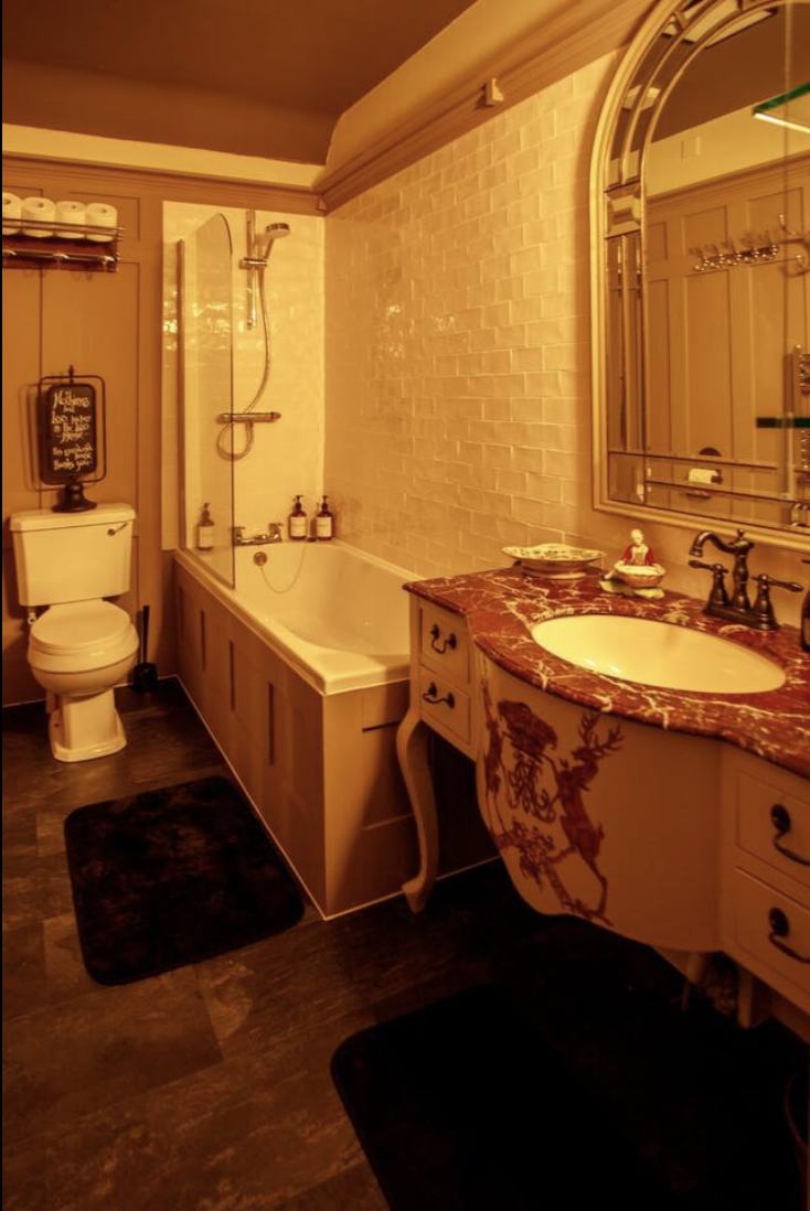 Bathroom with a toilet, bathtub with a glass shower door, marble vanity with a red and brown pattern, and a large mirror. The walls have brick-like tiles, and the floor is dark wood or wood-like material.