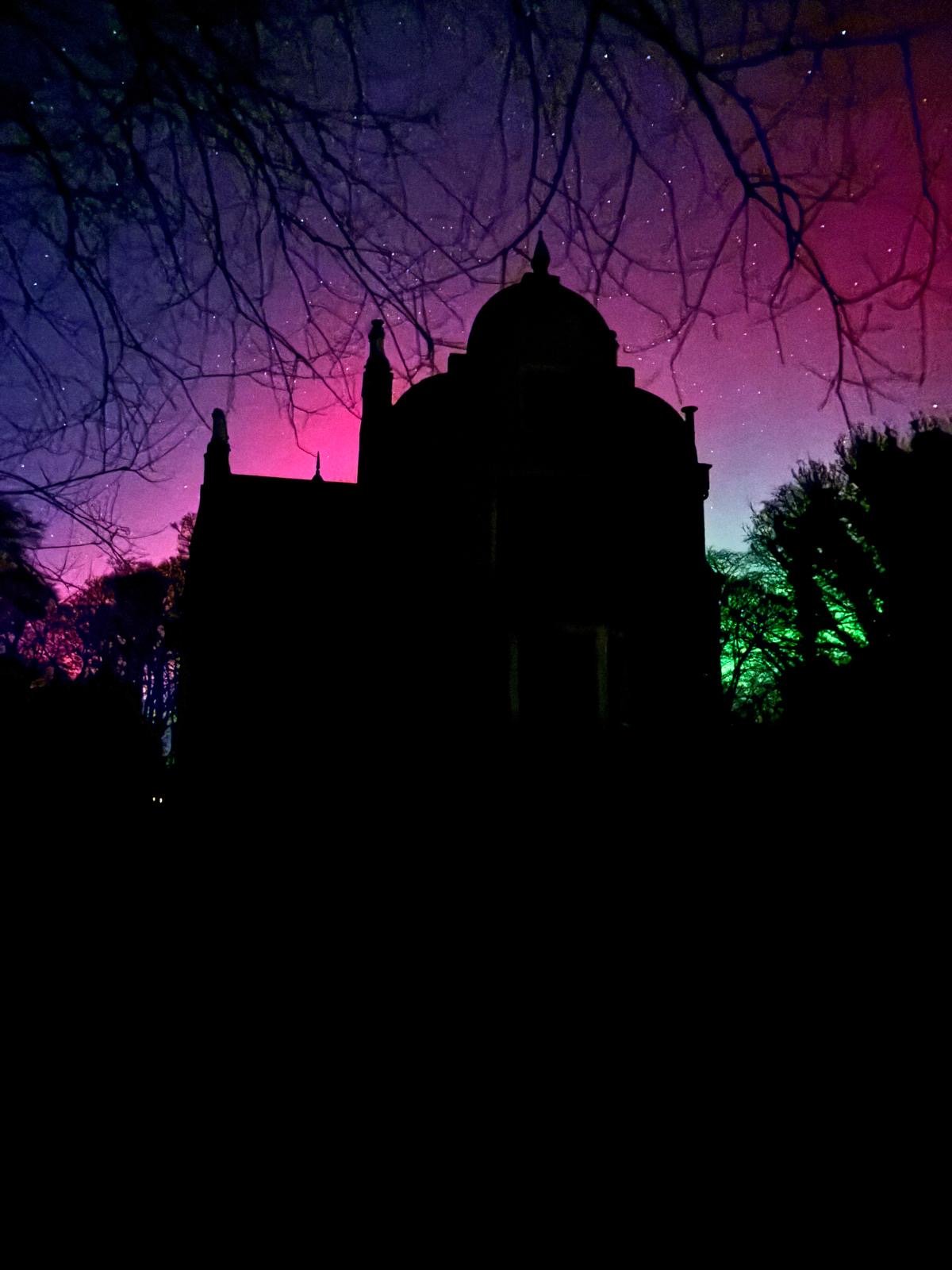 Silhouette of a building with a dome and spires, trees with bare branches, against a colorful night sky with stars and aurora borealis in pink, purple, and green hues.