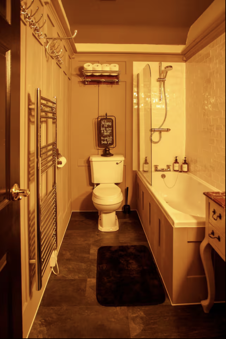 A cozy bathroom with a toilet, black mat, bathtub, and various toiletries on the ledge. Towel racks and rolls of paper towels are also visible.