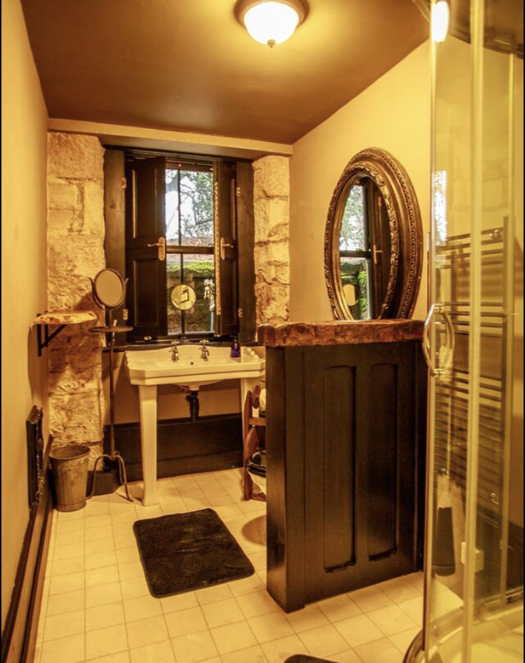 Bathroom with stone wall accents, a white sink with vintage fixtures, a large oval mirror, a window with shutters, a small shelf, and a black vanity, with a shower enclosure on the right.
