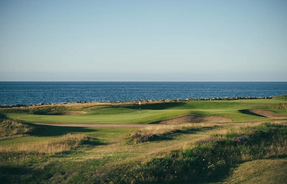 A golf course with green fairways and sand traps, overlooking the ocean under a clear sky.