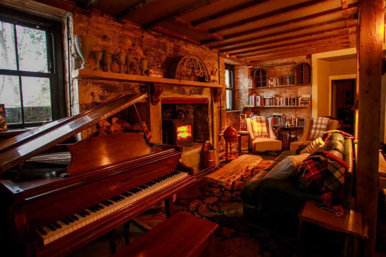 Cozy living room with a grand piano, stone fireplace with a fire, bookshelves filled with books, and comfy chairs with plaid blankets.