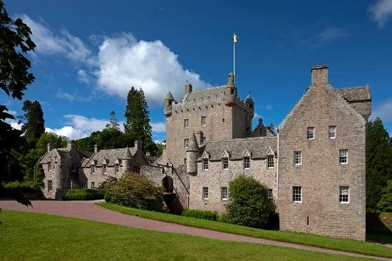 A stone castle with towers and a flag, surrounded by green grass and trees, under a partly cloudy sky.