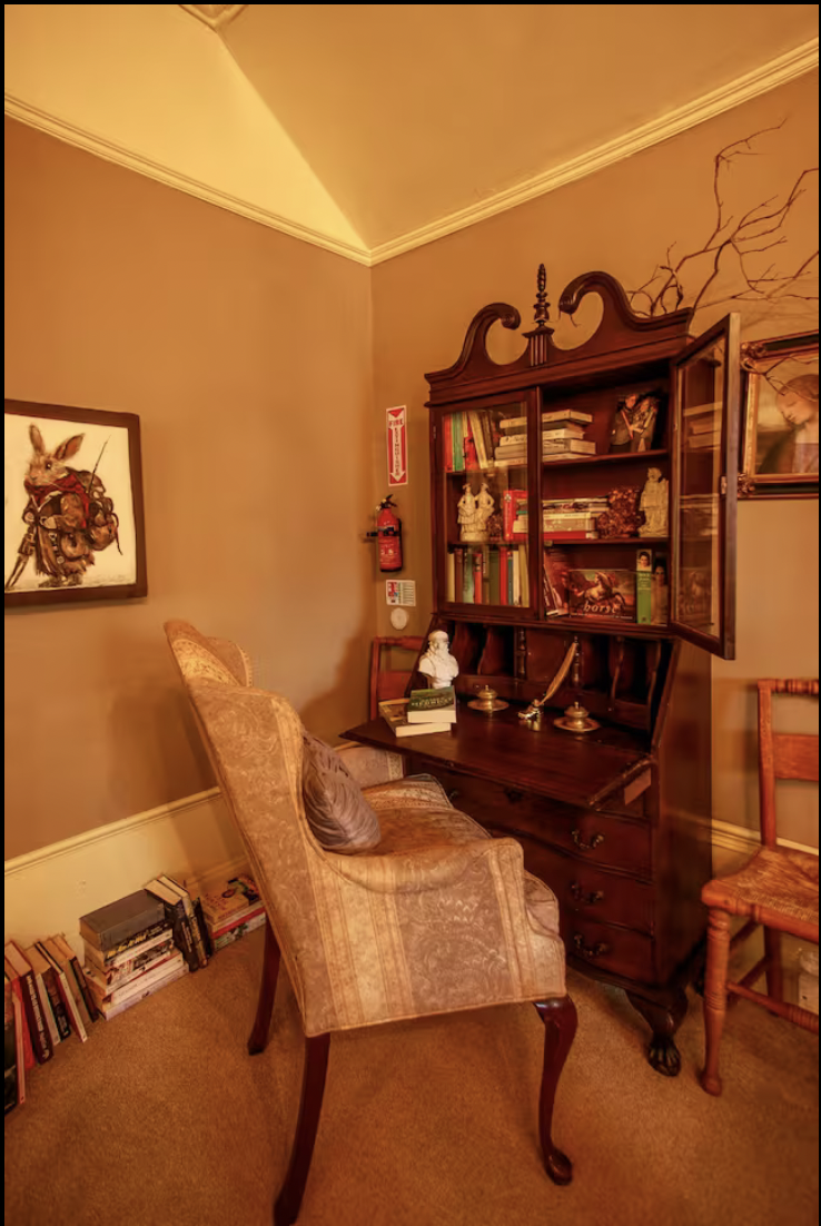 A cozy corner in a living room with a vintage armchair, a wooden bookshelf, and various decorative items. The corner also has a small side table and a framed bunny artwork on the wall.