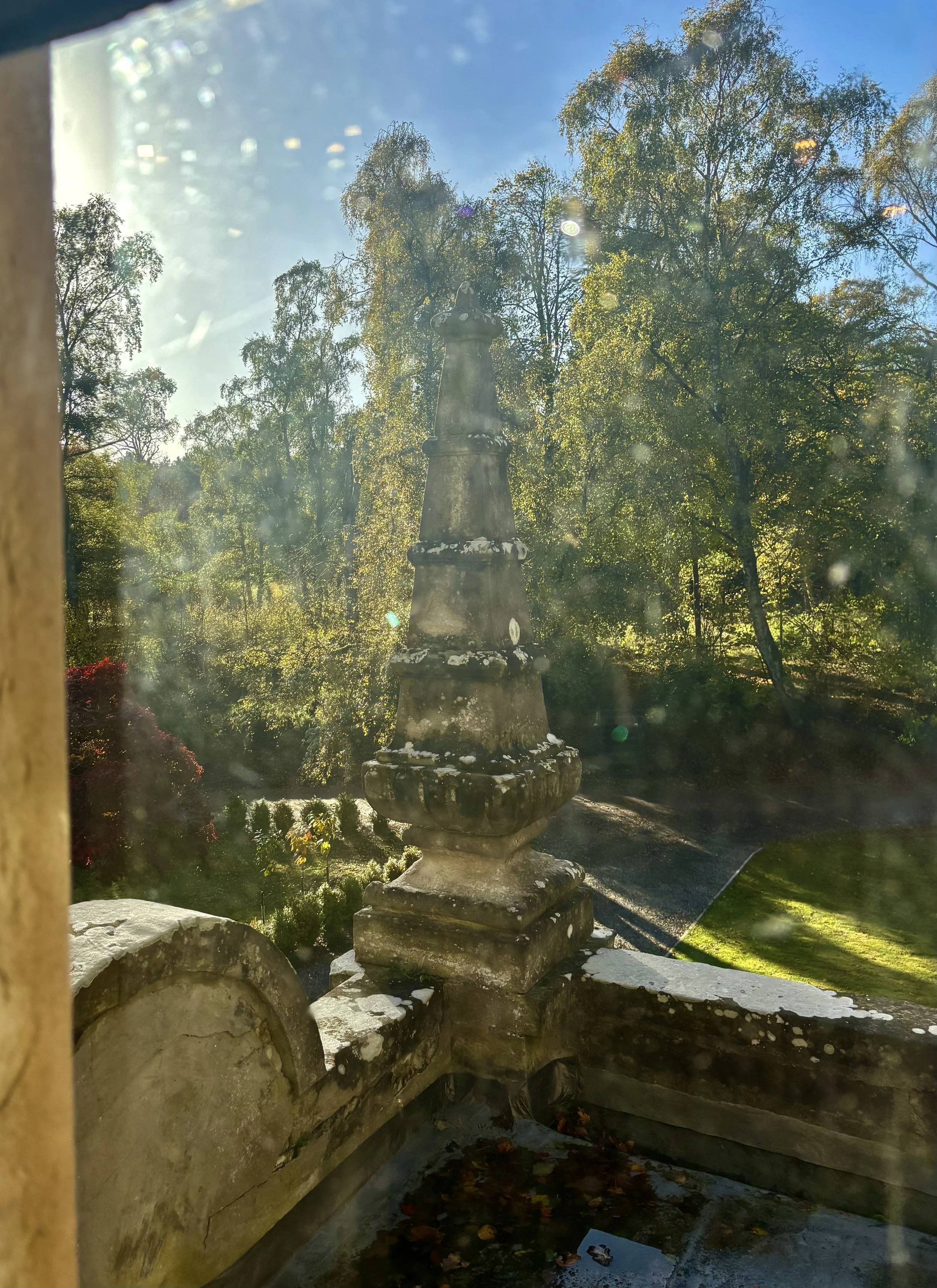 View of an outdoor stone monument or pagoda seen through a window with some dirt and smudges, surrounded by trees with green leaves, under a bright blue sky.