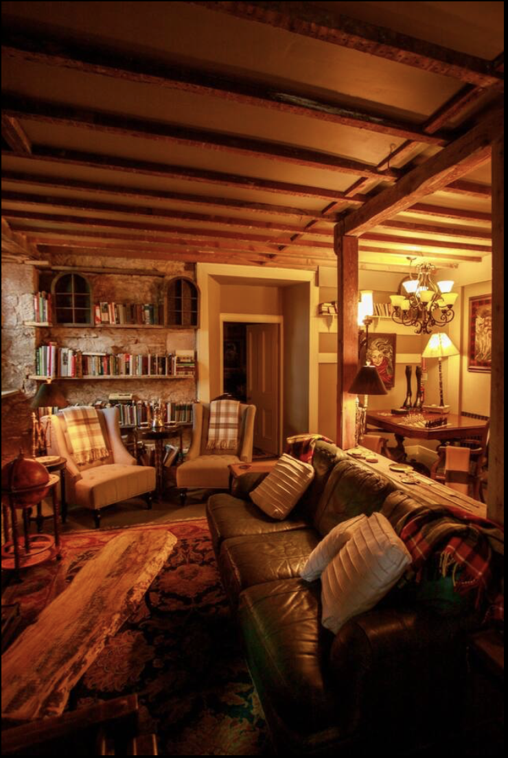 Cozy living room with leather sofa, plaid pillows, wooden coffee table, armchairs with striped cushions, bookshelf, and a dining area with chandelier lighting.