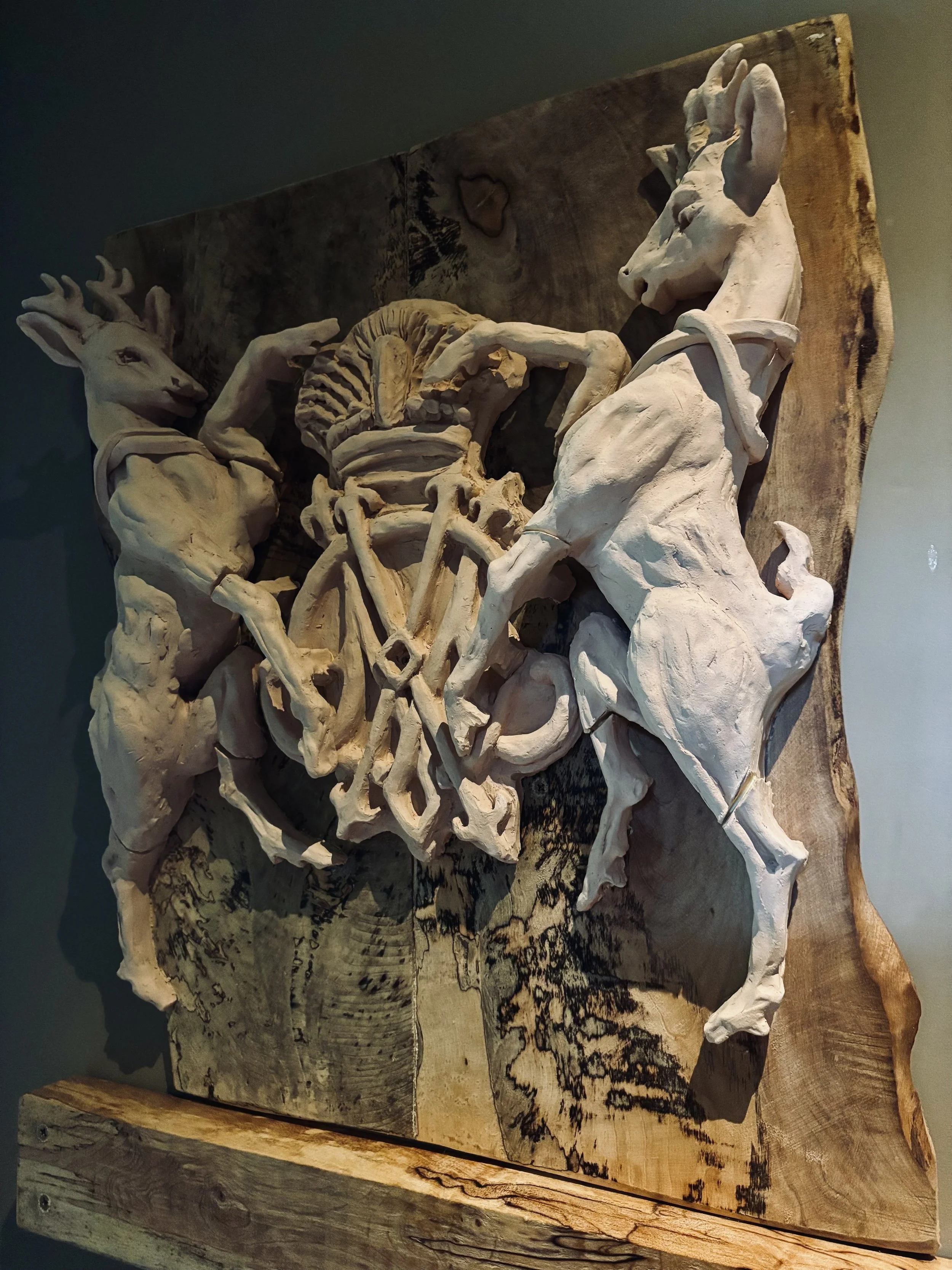 A carved wooden sculpture of two humanoid animals with elongated heads and limbs, holding a wheel, displayed on a textured wooden background.