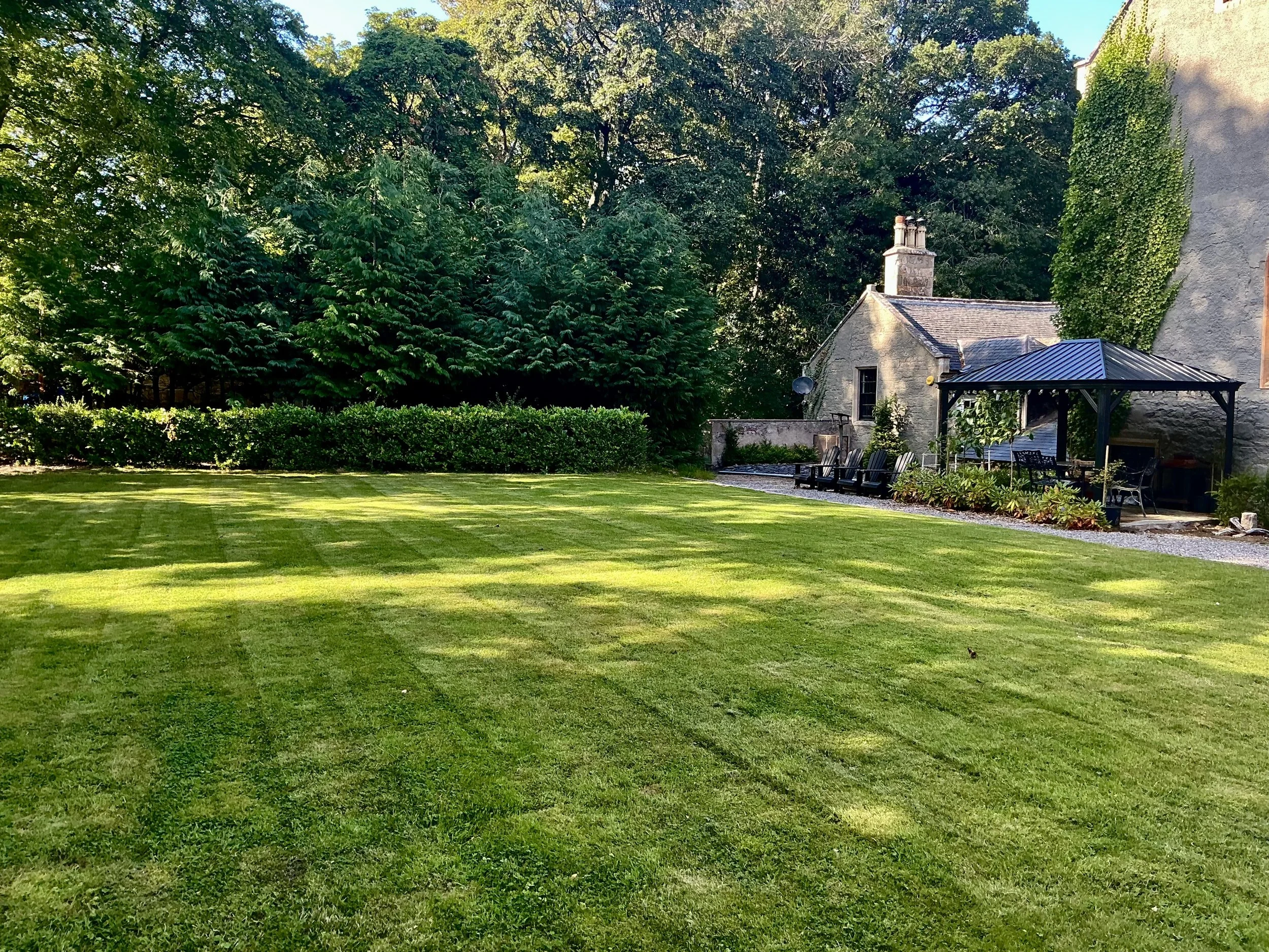 A backyard lawn with neatly mowed grass, bordered by shrubs and trees, featuring a stone house with a chimney, a covered seating area, and outdoor chairs.