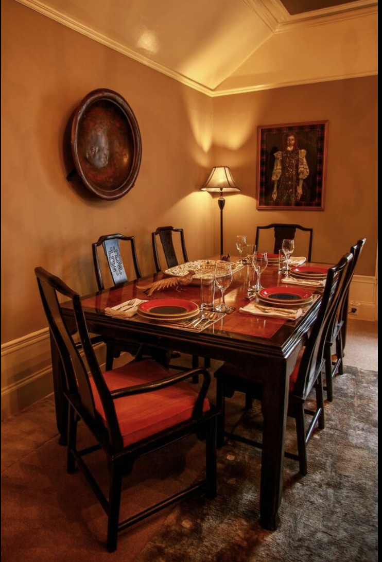 A dining room with a wooden table set for six, with red and black plates, glasses, napkins, and utensils. There are six chairs, a floor lamp, and wall decorations including a framed portrait of a woman and a large round artwork. The room has warm lig
