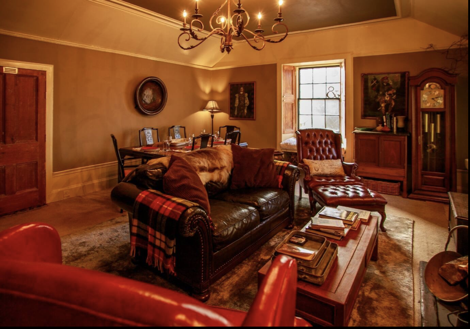 Cozy living room with a leather sofa, plaid blanket, and pillows, illuminated by a chandelier and a floor lamp, featuring a window with wooden shutters, a wooden coffee table with books, and antique furniture and artwork.