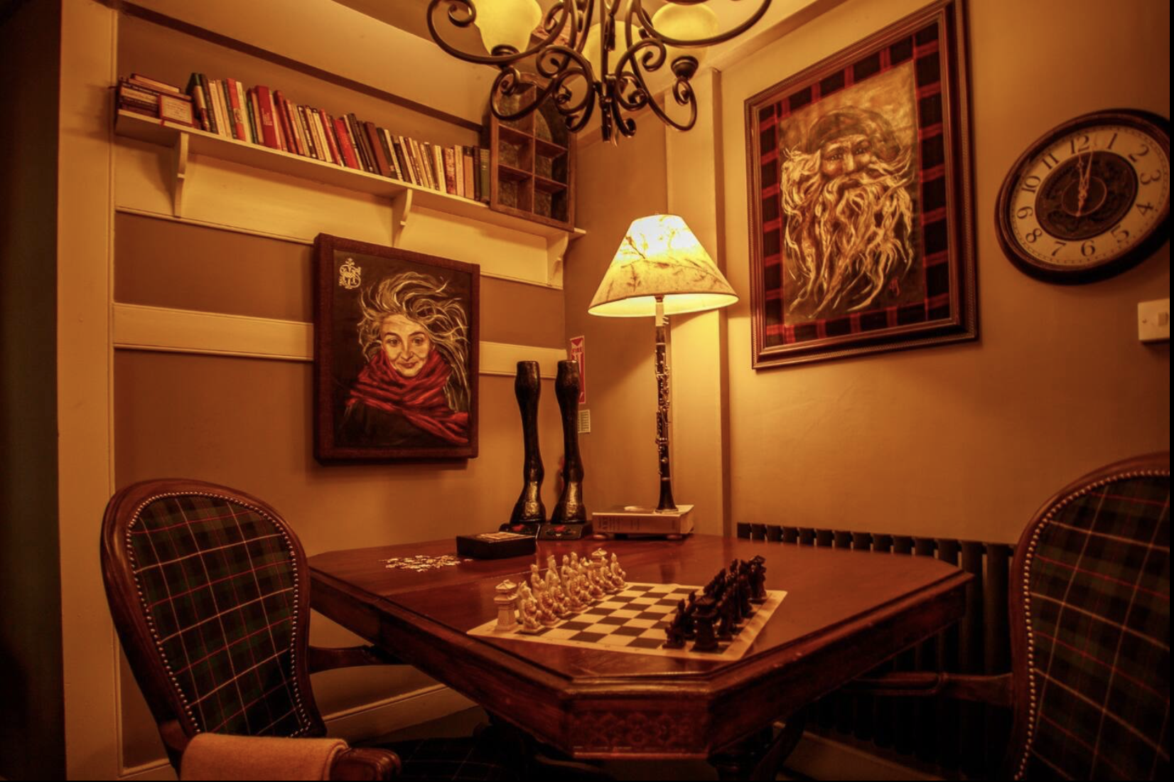 A cozy room with a chess set on a wooden table, plaid upholstered chairs, a bookshelf on the wall, framed artwork of a woman and a bearded man, a lamp, and a clock showing 12:00.