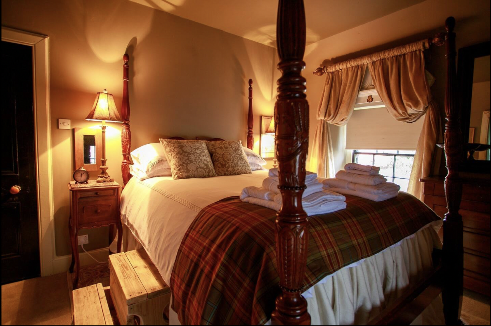 Cozy bedroom with four-poster bed, folded towels on bed, wooden bedside tables with lamps, window with curtains, and warm lighting.