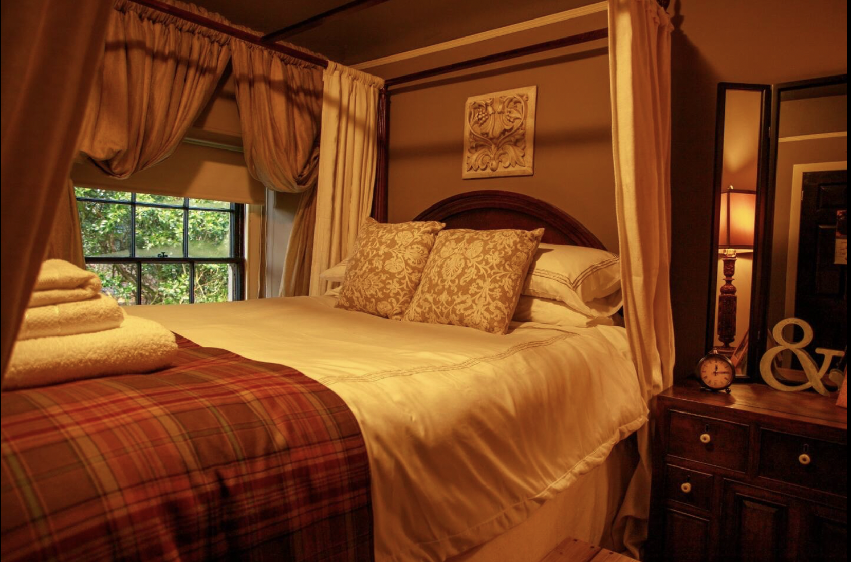 Cozy bedroom with a four-poster bed, gold and cream bedding, plaid blanket, closed window with lush greenery outside, bedside table with warm lighting, decorative wall art, and folded towels.