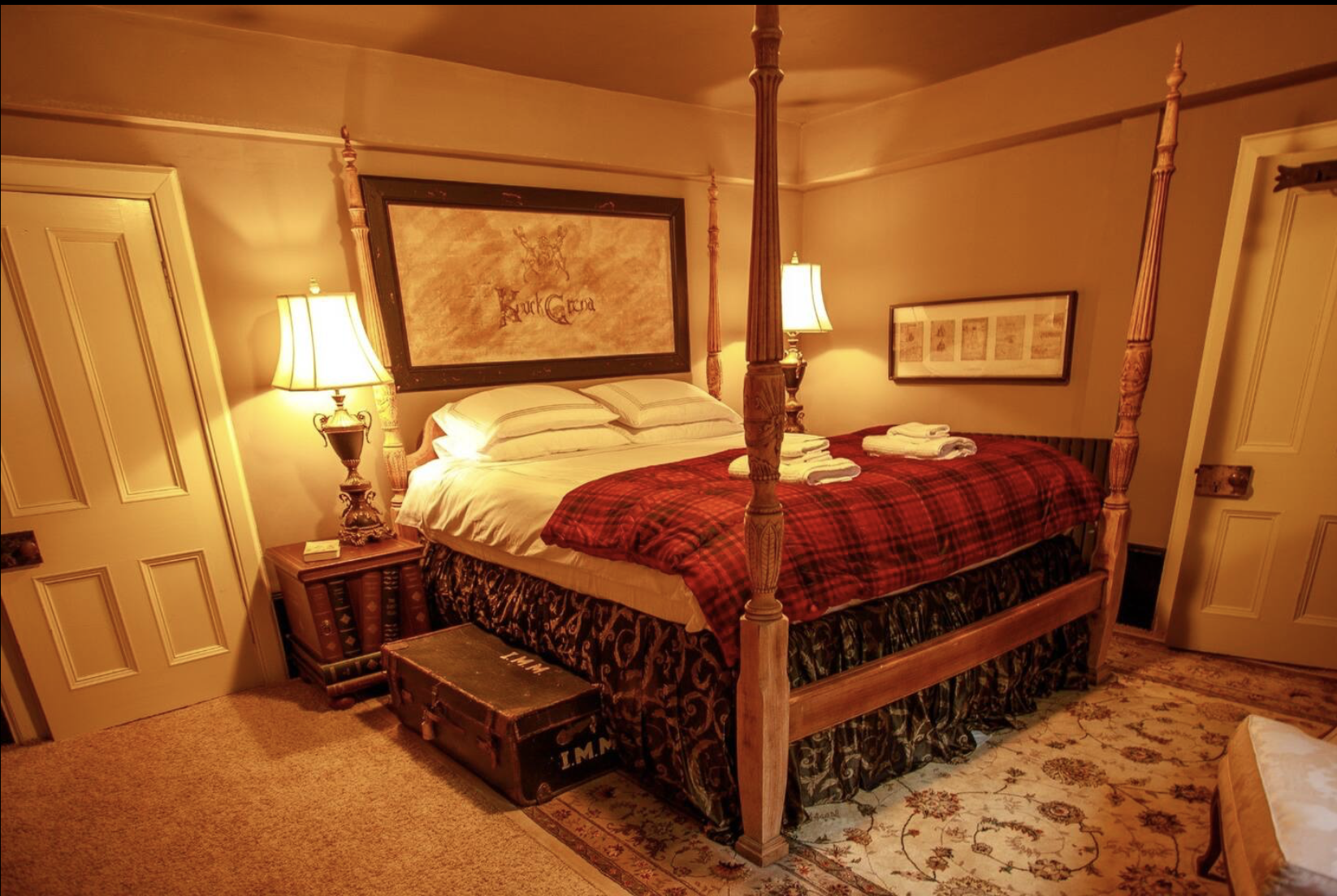A cozy bedroom with a four-poster wooden bed, neatly made, with a red plaid blanket and folded towels on top. There are two nightstands with lamps on each side and framed artwork on the walls. A vintage trunk is at the foot of the bed, and the room h