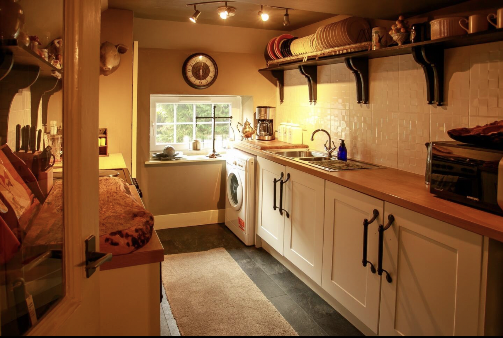Cozy kitchen with white cabinets, wooden countertop, and wall-mounted shelves holding dishes, oven mitts, and kitchenware. Small window above the sink shows greenery outside. Various kitchen appliances are on the counters, and a washing machine is pl