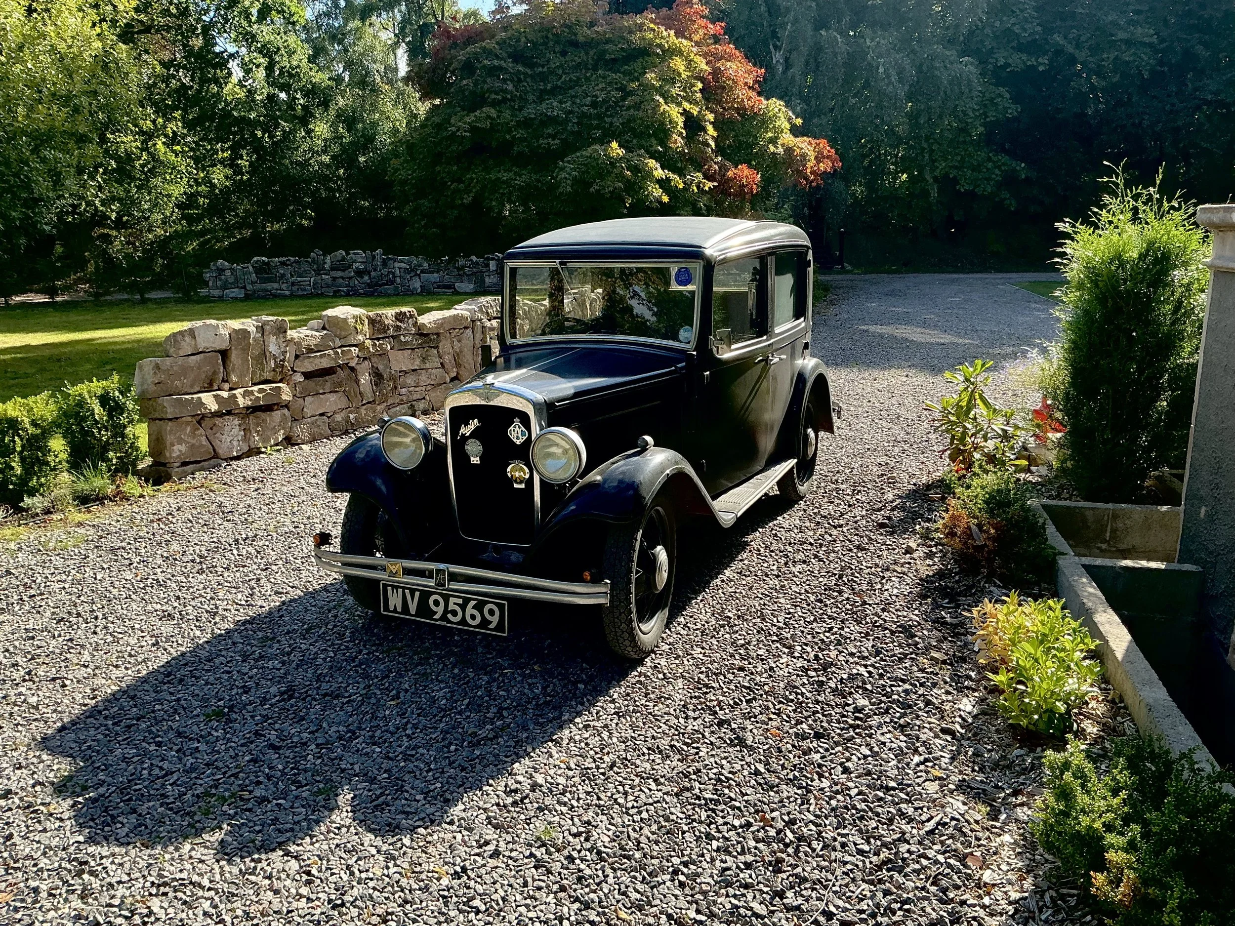 A vintage black car parked on a gravel driveway with a stone wall and green trees in the background.