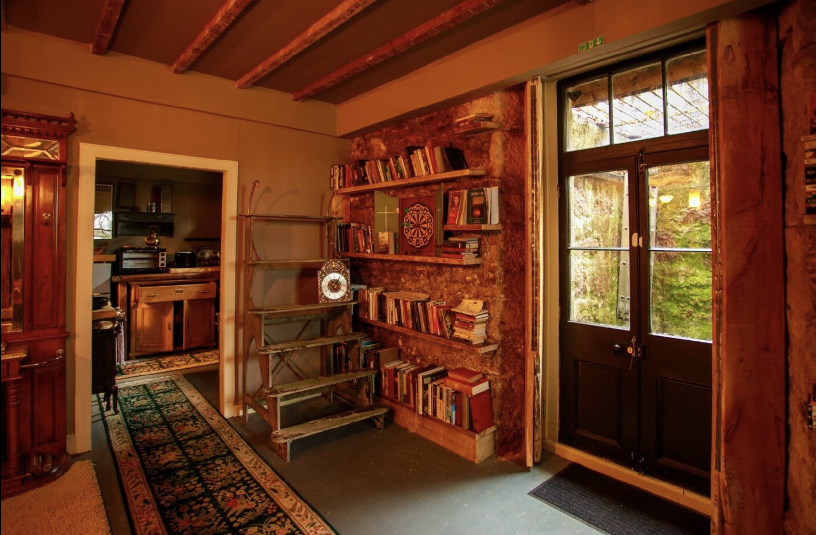 Interior view of a cozy room with brick walls, a black door with glass panes, and bookshelves filled with books. There's a doorway leading to another room with wooden furniture and a window. The room has a patterned rug and exposed wooden ceiling bea