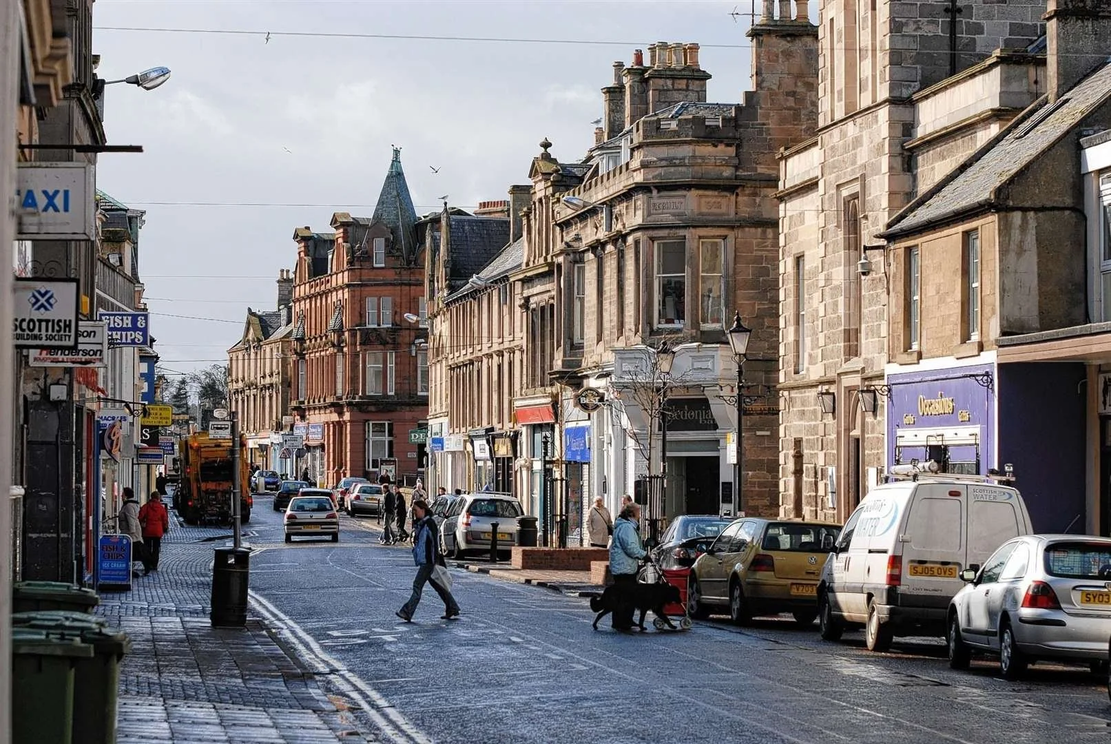 A busy city street with pedestrians walking across the crosswalk, parked cars, and buildings with shops and offices. Overcast sky with some birds flying overhead.