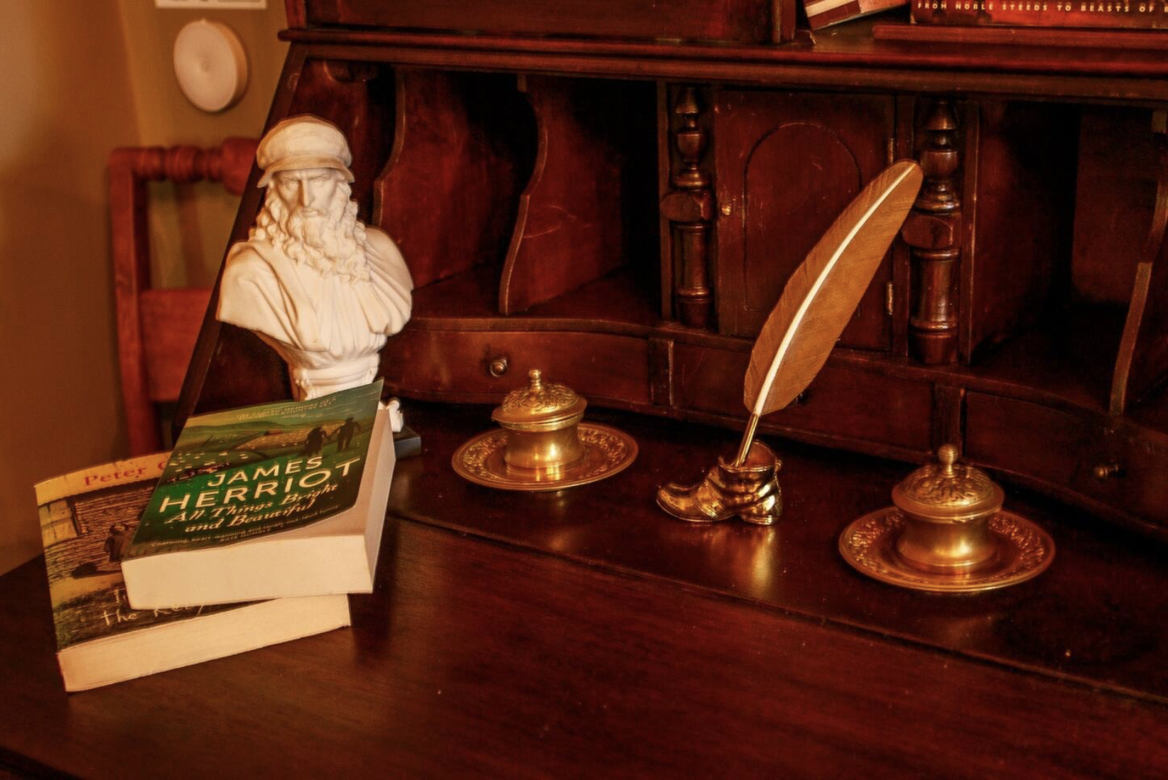 A wooden desk with a small bust of a bearded man wearing a cap, a gold inkwell with a matching plate, a large feather quill in a gold holder, and a large paperback book titled 'James Herriot: All Creatures Bright and Beautiful'.