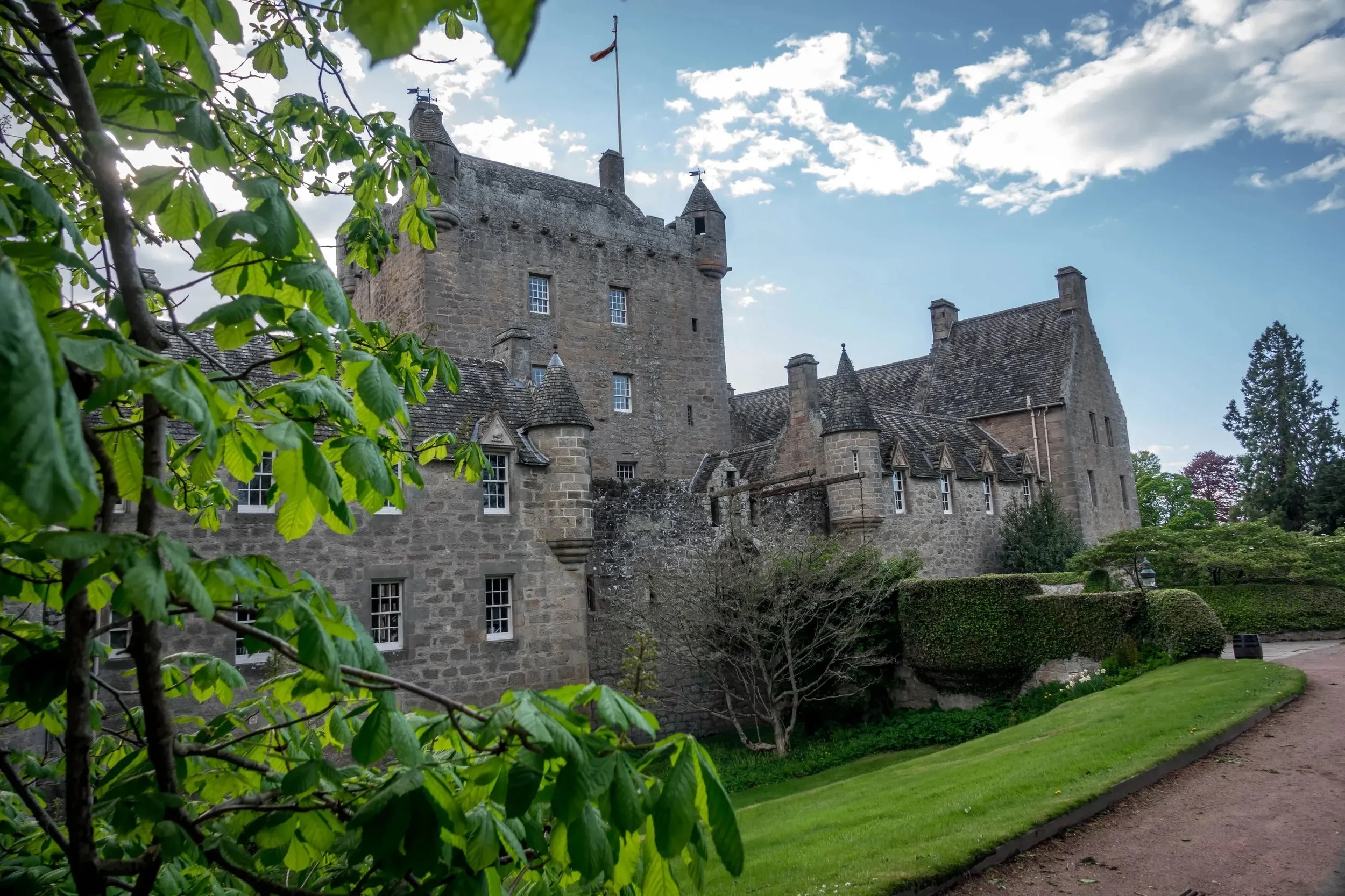 A historic stone castle with turrets and small windows, surrounded by green trees and bushes, under a partly cloudy sky with sunlight