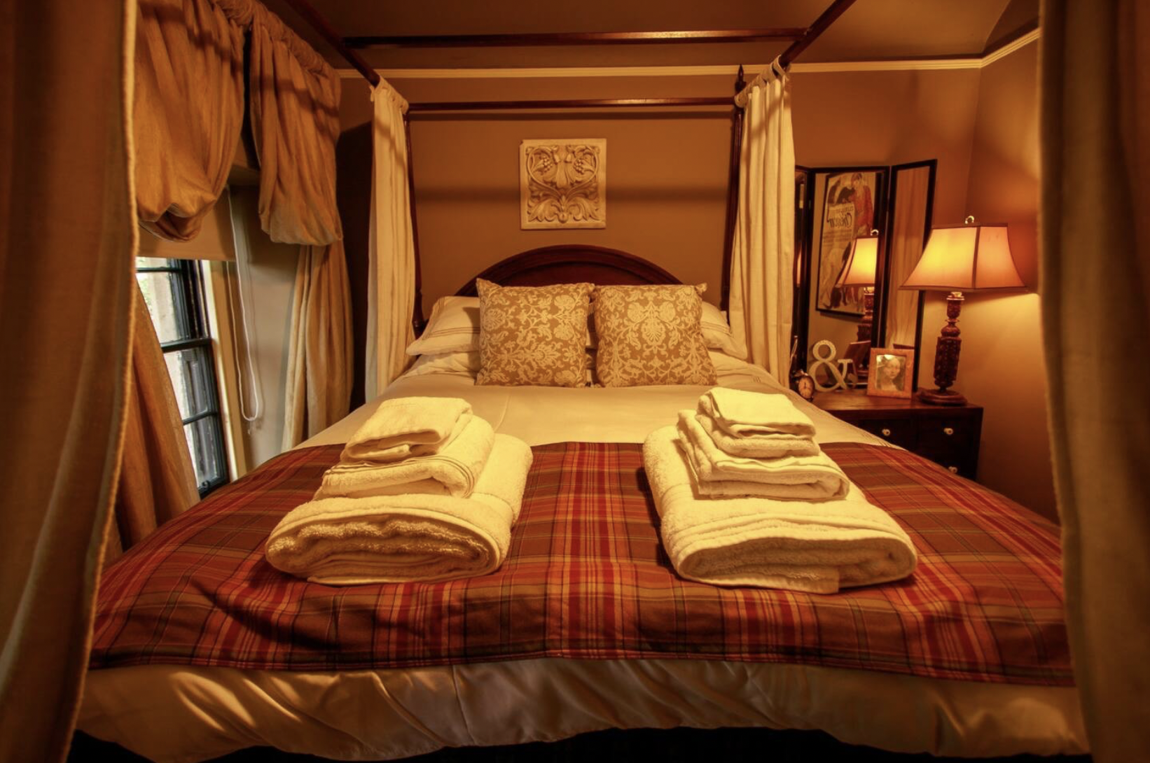A cozy bedroom with a four-poster bed draped in white curtains, layered with white and beige pillows, and topped with folded towels on a plaid bedspread. A nightstand with a lit lamp, framed pictures, and decorative items is beside it. Warm lighting 