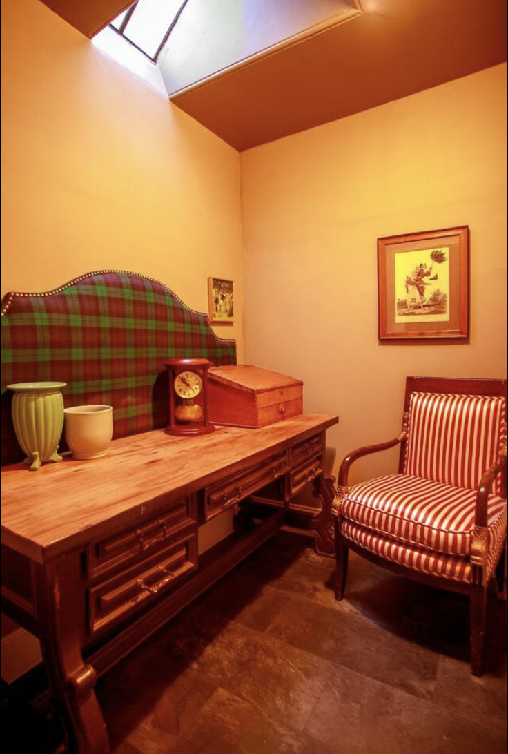 Corner of a room with a wooden table, a striped armchair, two framed pictures on the wall, and a small window near the ceiling.
