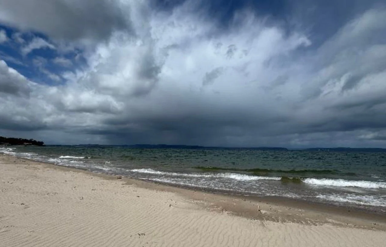Cloudy sky over a sandy beach with small waves, distant shoreline.