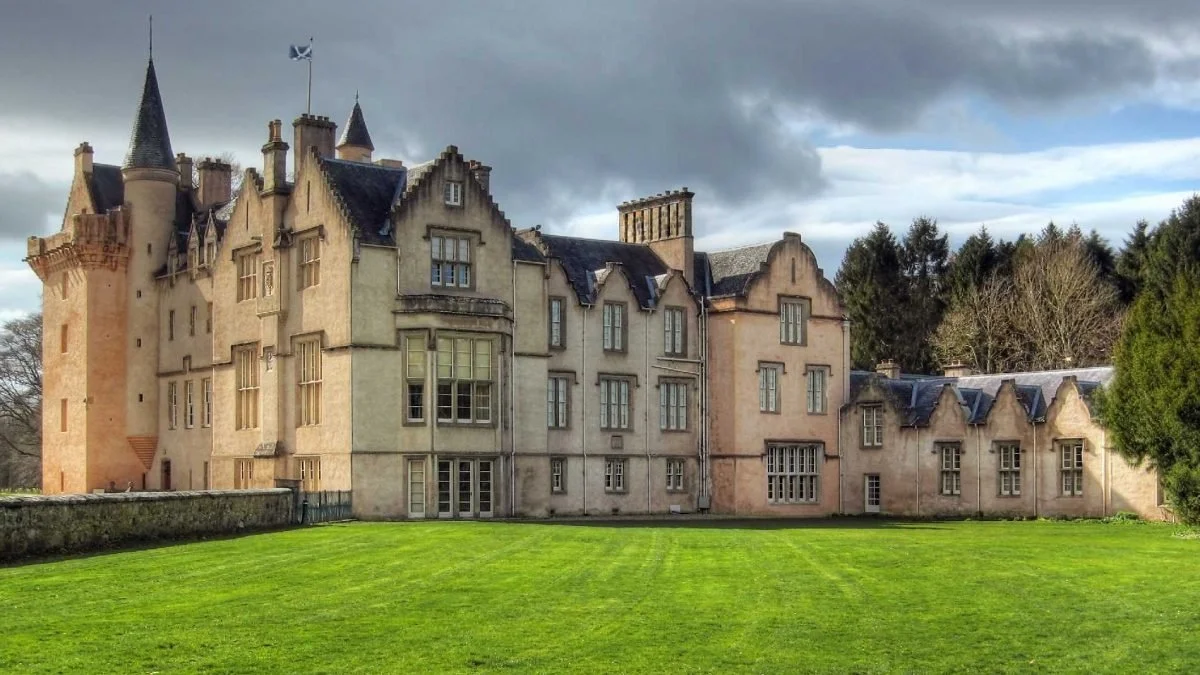 A large historic castle with tall towers and turrets, surrounded by a well-maintained green lawn and trees, under a cloudy sky.