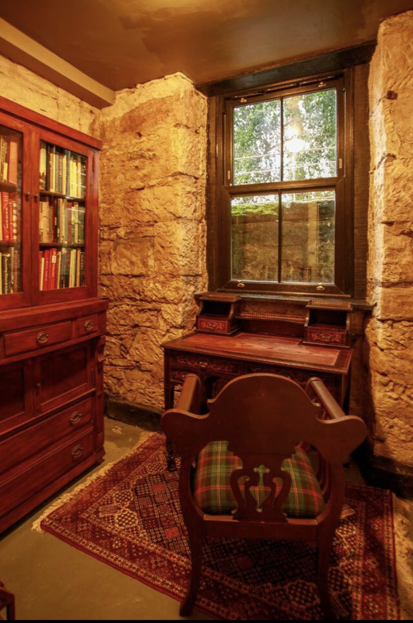 A cozy room with rustic stone walls, a large window showing greenery outside, a wooden desk, a wooden chair with a plaid cushion, a bookshelf filled with books, and a patterned area rug.