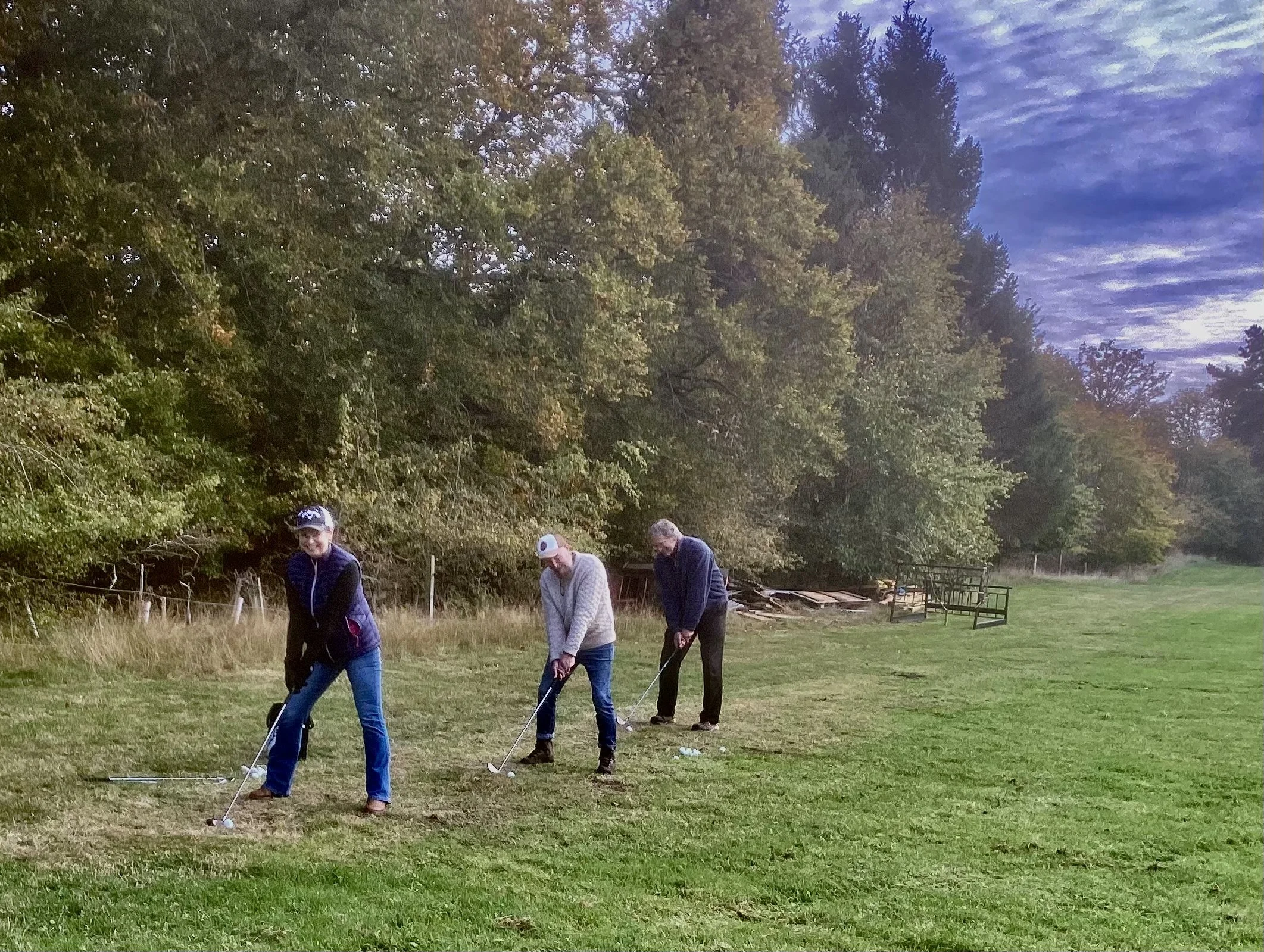 Three people playing golf on a grassy field with trees in the background during early evening.