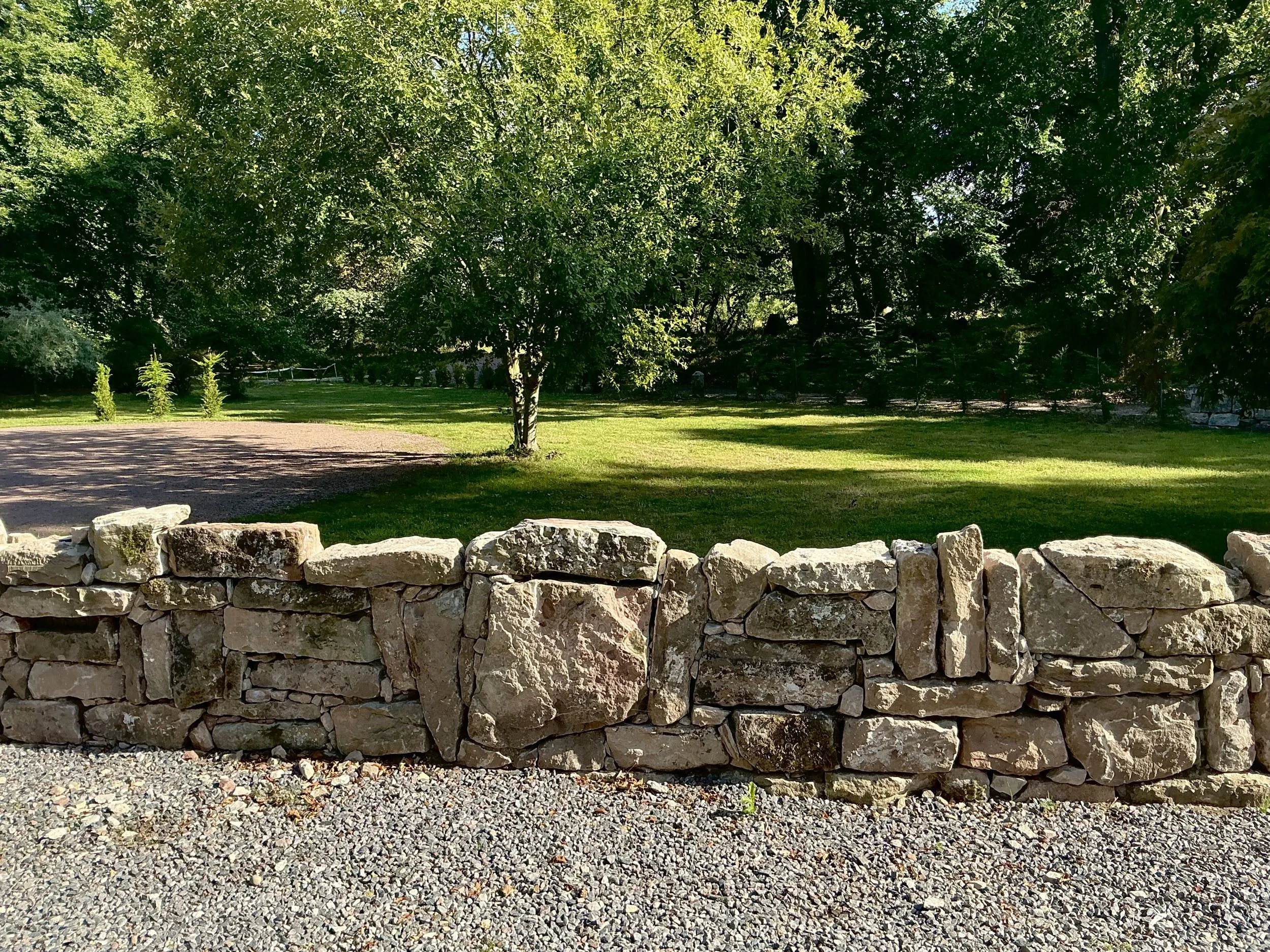A stone wall in the foreground with a gravel path in front and lush green trees and grass in the background, sunny day.