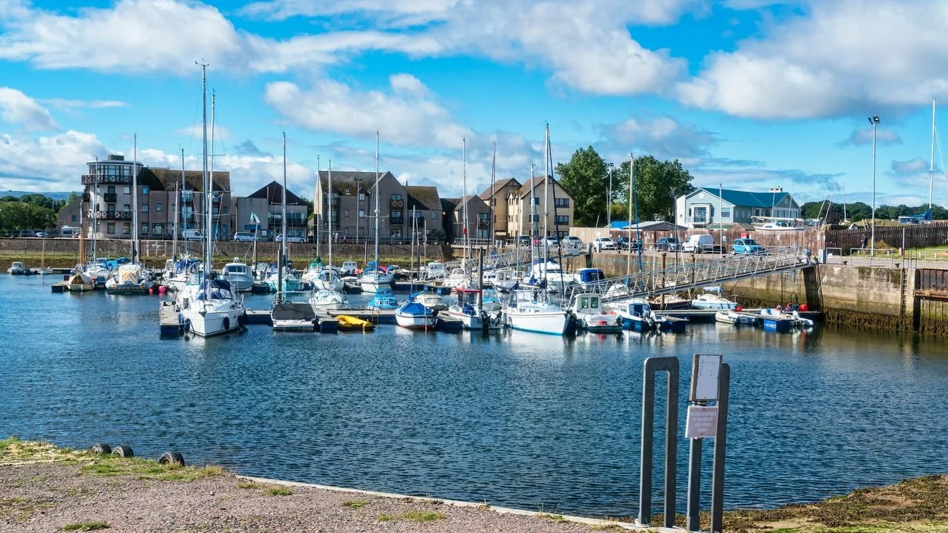 A marina with several sailboats and motorboats docked, buildings and houses in the background, and a partly cloudy sky overhead.