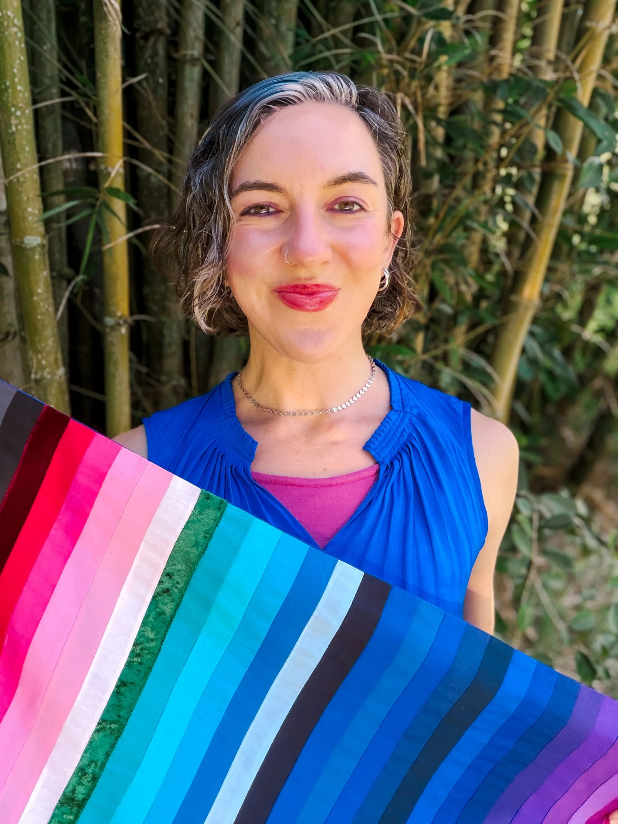 A woman with short curly hair, wearing a blue top, smiling outdoors in front of bamboo plants, holding a rainbow-colored fabric or flag.