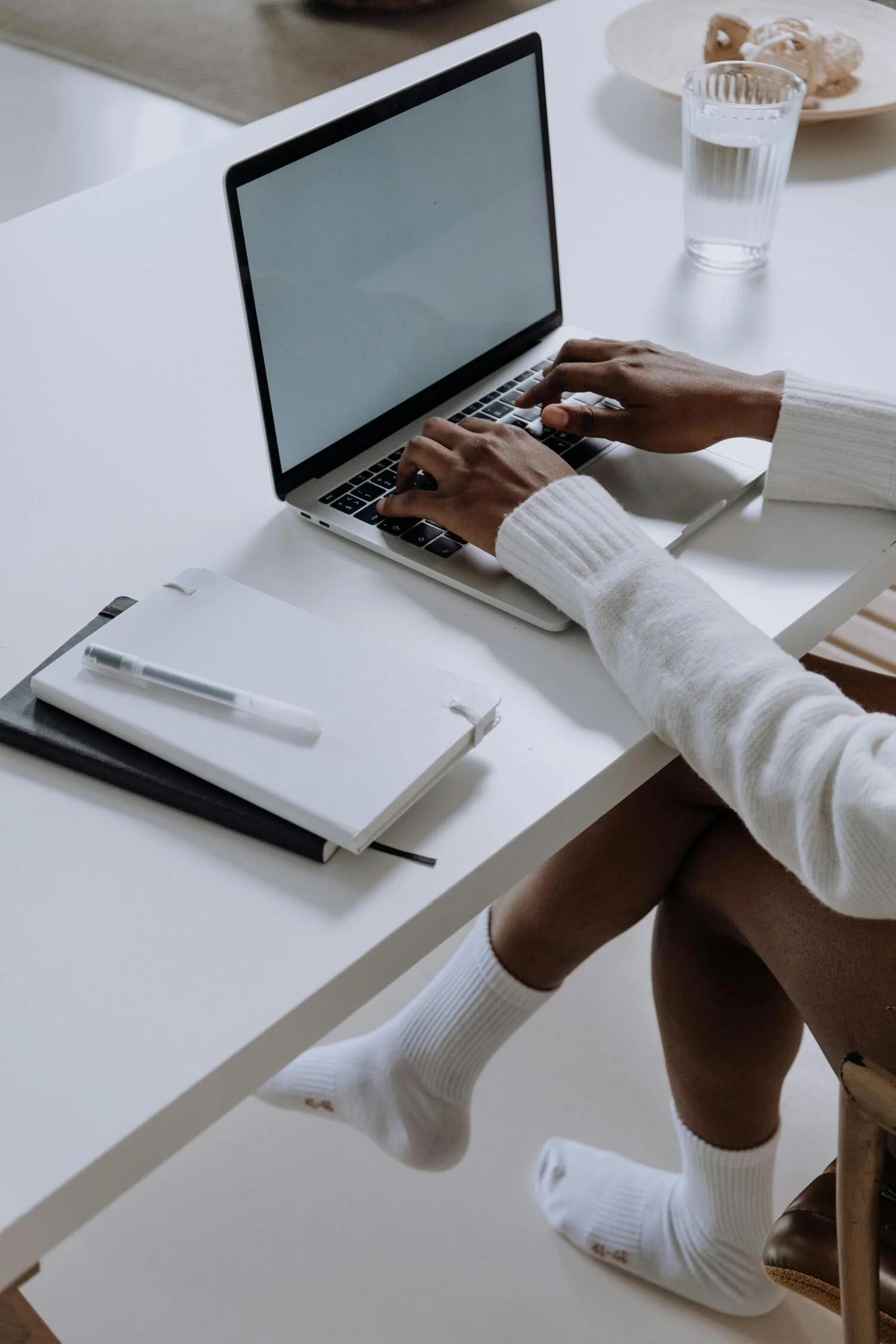 Person wearing white socks and brown boots sitting at a white table with a laptop, a notebook with a pen, a glass of water, and a plate with snacks