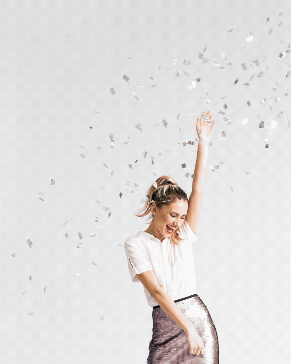 A woman in a white shirt and shiny skirt celebrating with her arm raised while confetti falls around her.