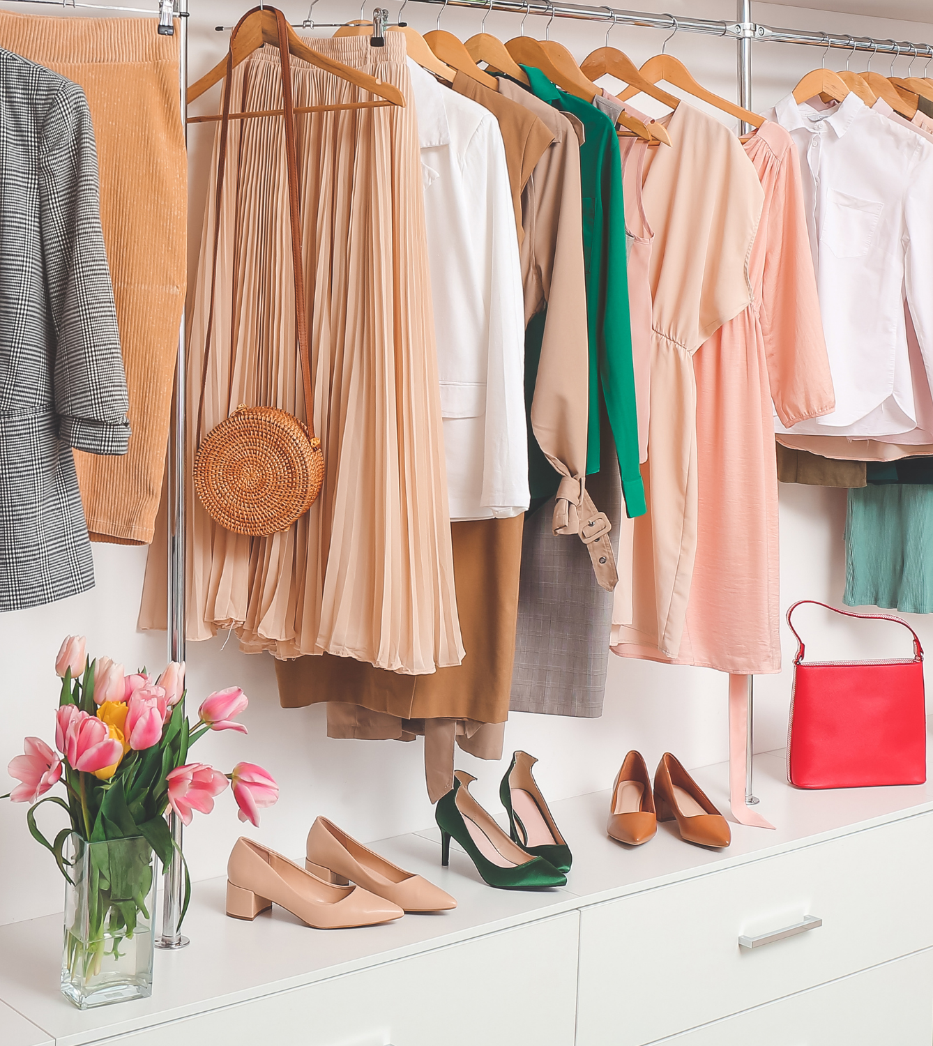 A display of women's clothing and accessories including dresses, blouses, jackets, shoes, a red handbag, and a bouquet of pink tulips on a white shelf with a clothing rack in the background.